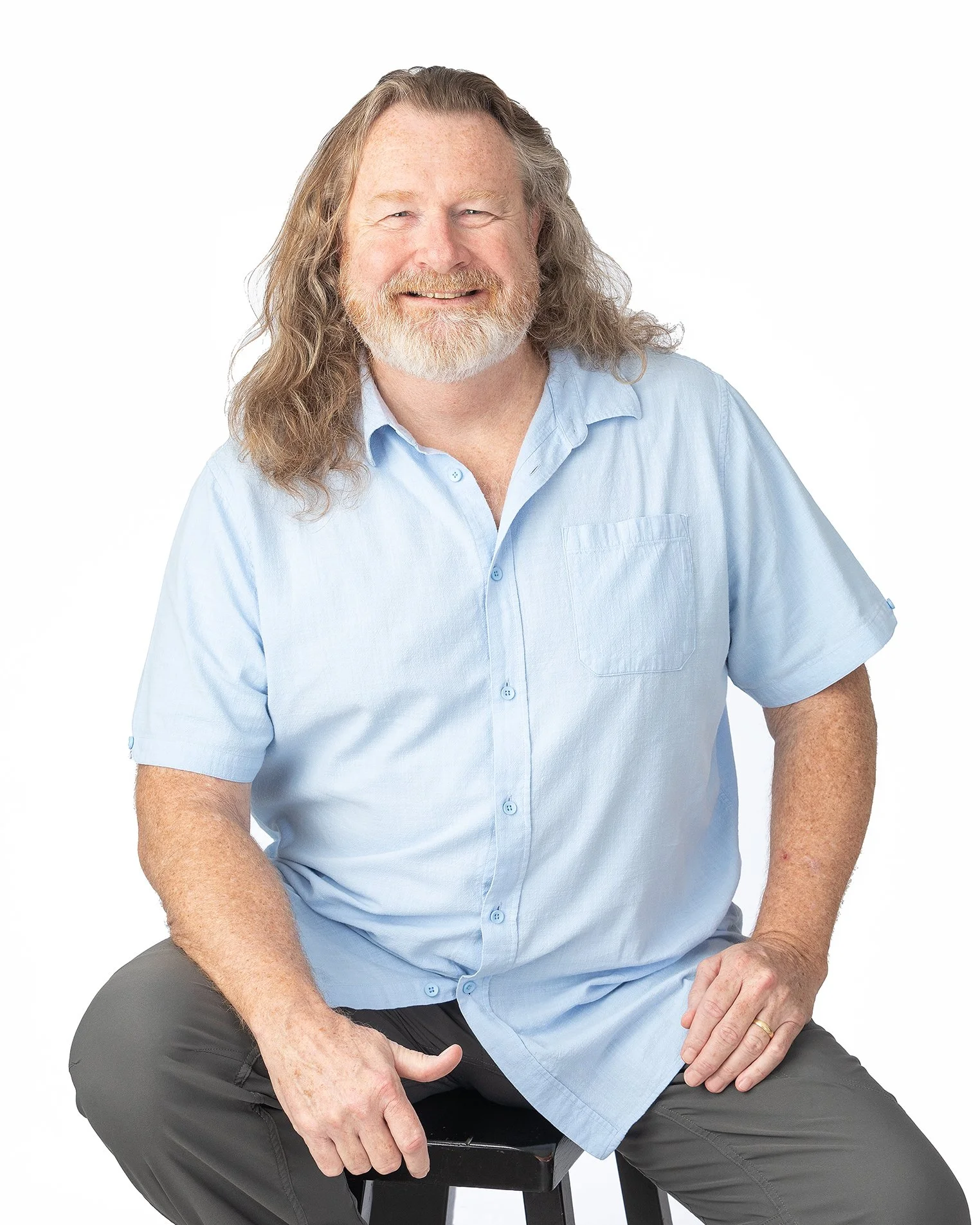 A smiling middle-aged man with long gray hair and a beard wearing a light blue short-sleeve button-up shirt and gray pants, sitting on a stool against a white background.