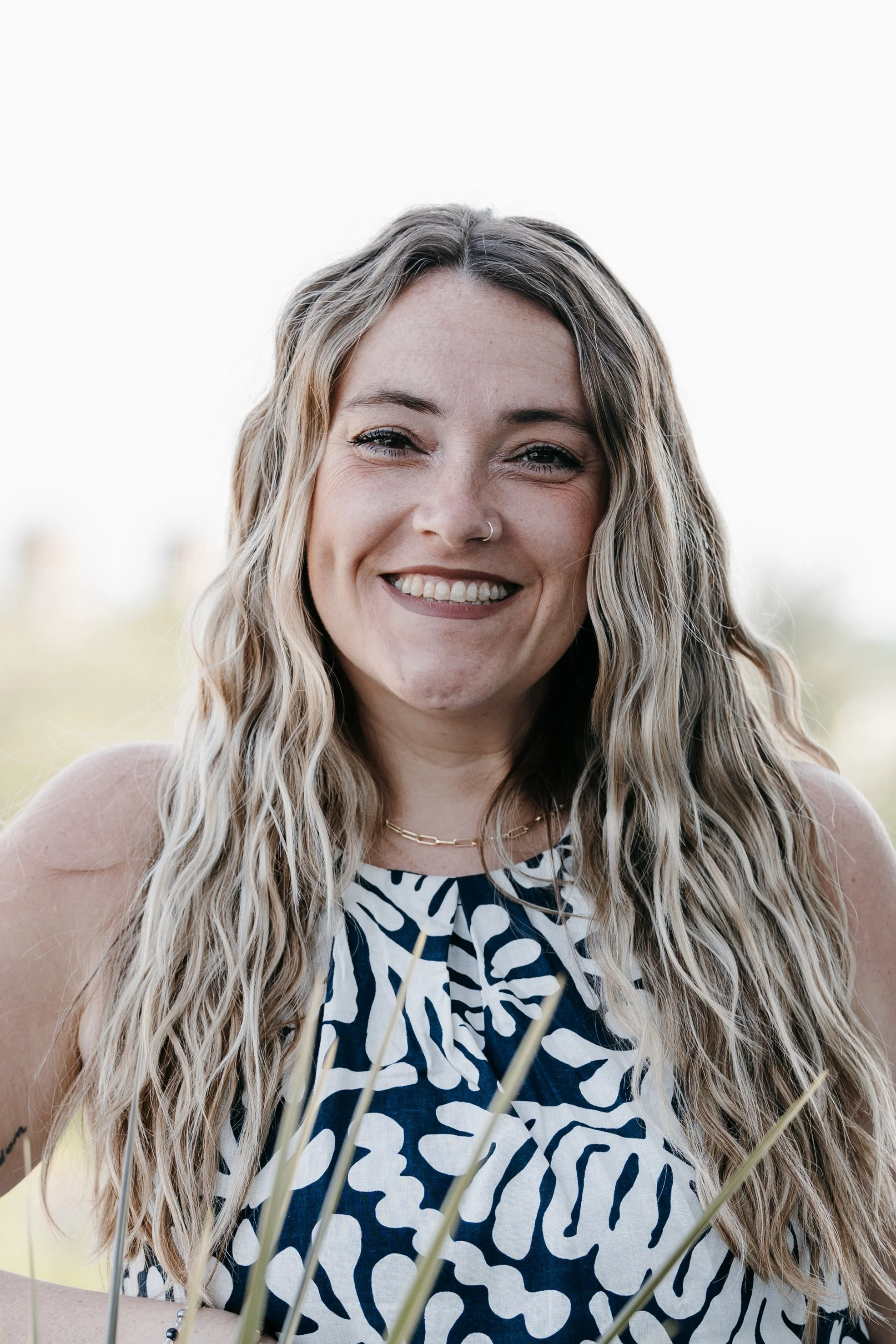 A smiling woman with long wavy blonde hair, wearing a navy and white patterned sleeveless top, a gold chain necklace, and a nose piercing, outdoors with a blurred background.