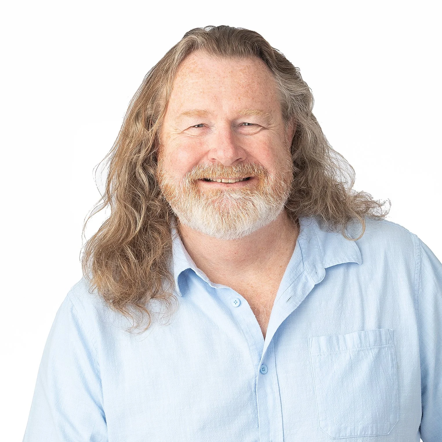 A smiling middle-aged man with long hair, a beard, and a mustache, wearing a light blue button-up shirt against a white background.