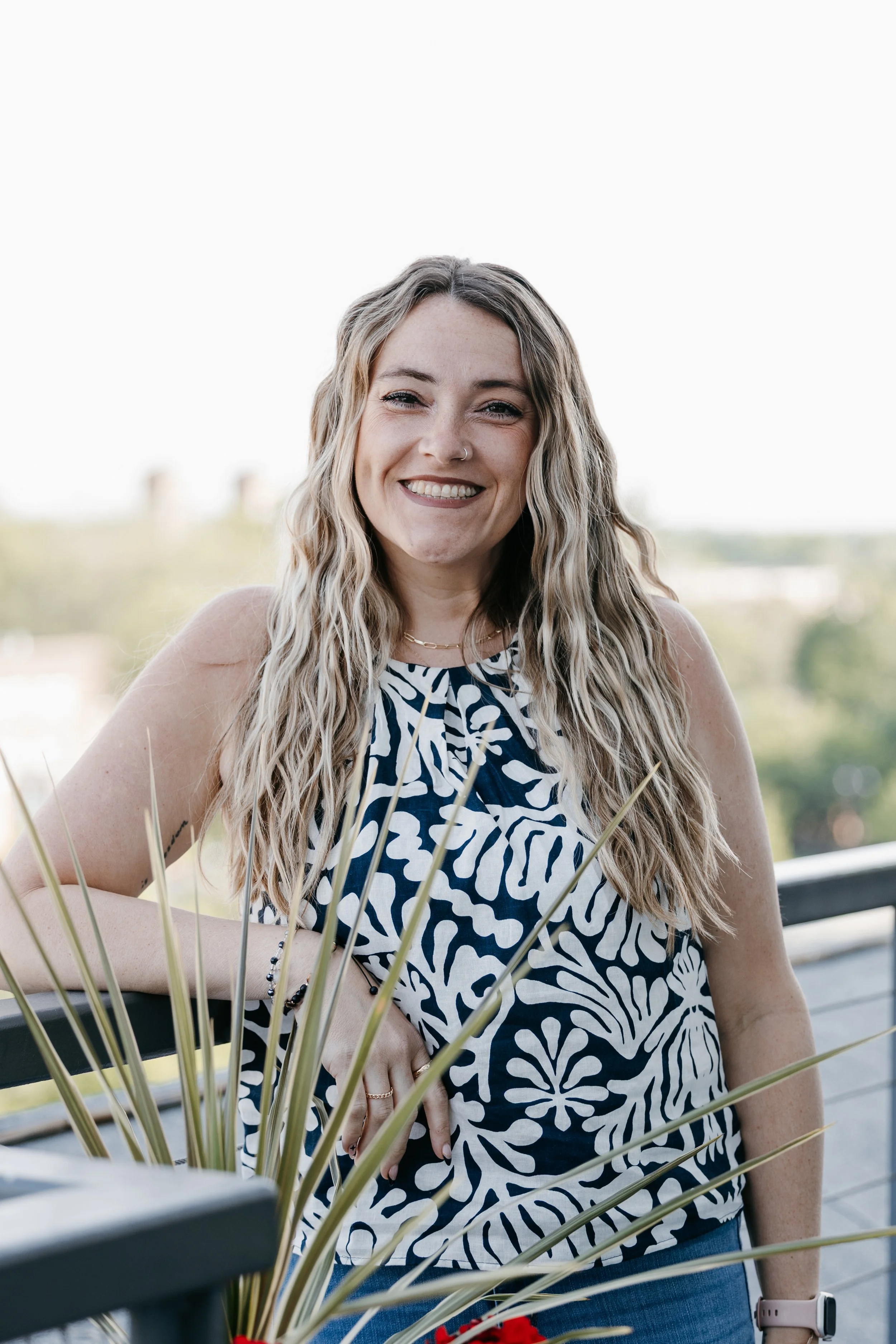 A smiling woman with blonde hair and a nose ring outdoors, wearing a white sleeveless top, a black beaded bracelet, and a smartwatch, with a blurred background of trees and a cityscape.
