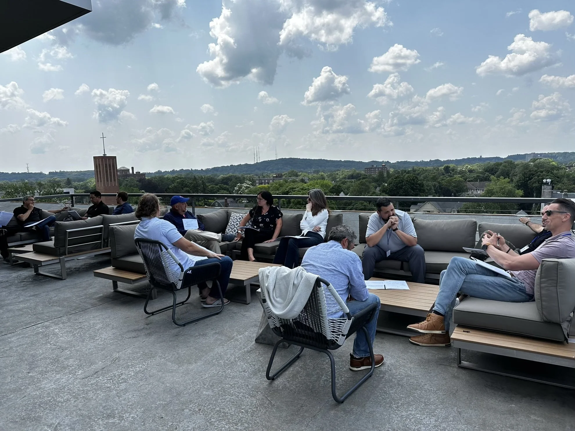 Group of people sitting and chatting on a rooftop terrace with a view of trees and hills under a partly cloudy sky.