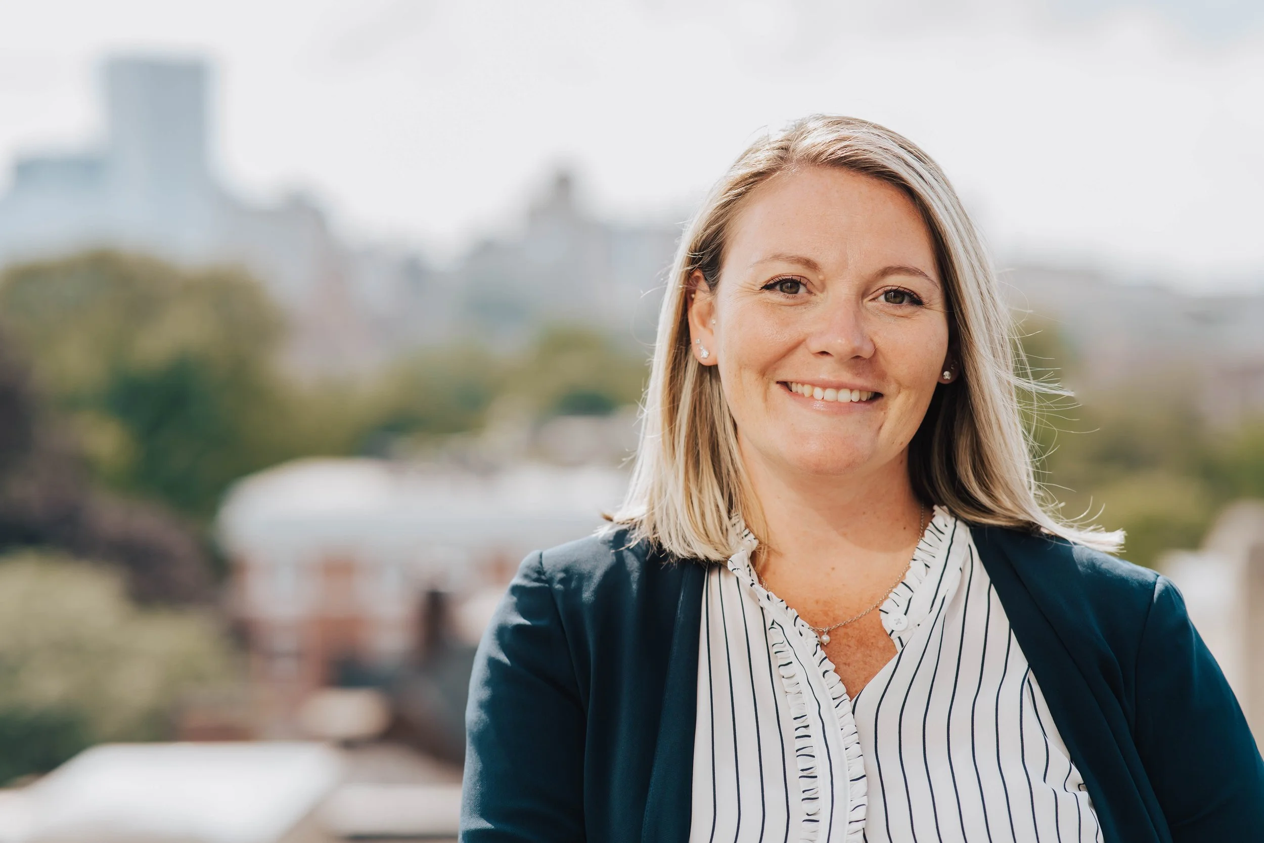 Smiling woman with shoulder-length blonde hair wearing a dark blazer and striped blouse, standing outdoors with blurred trees and cityscape background.