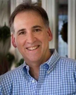 A smiling middle-aged man with short brown hair, wearing a blue checkered button-up shirt, standing indoors with a window and houseplant in the background.