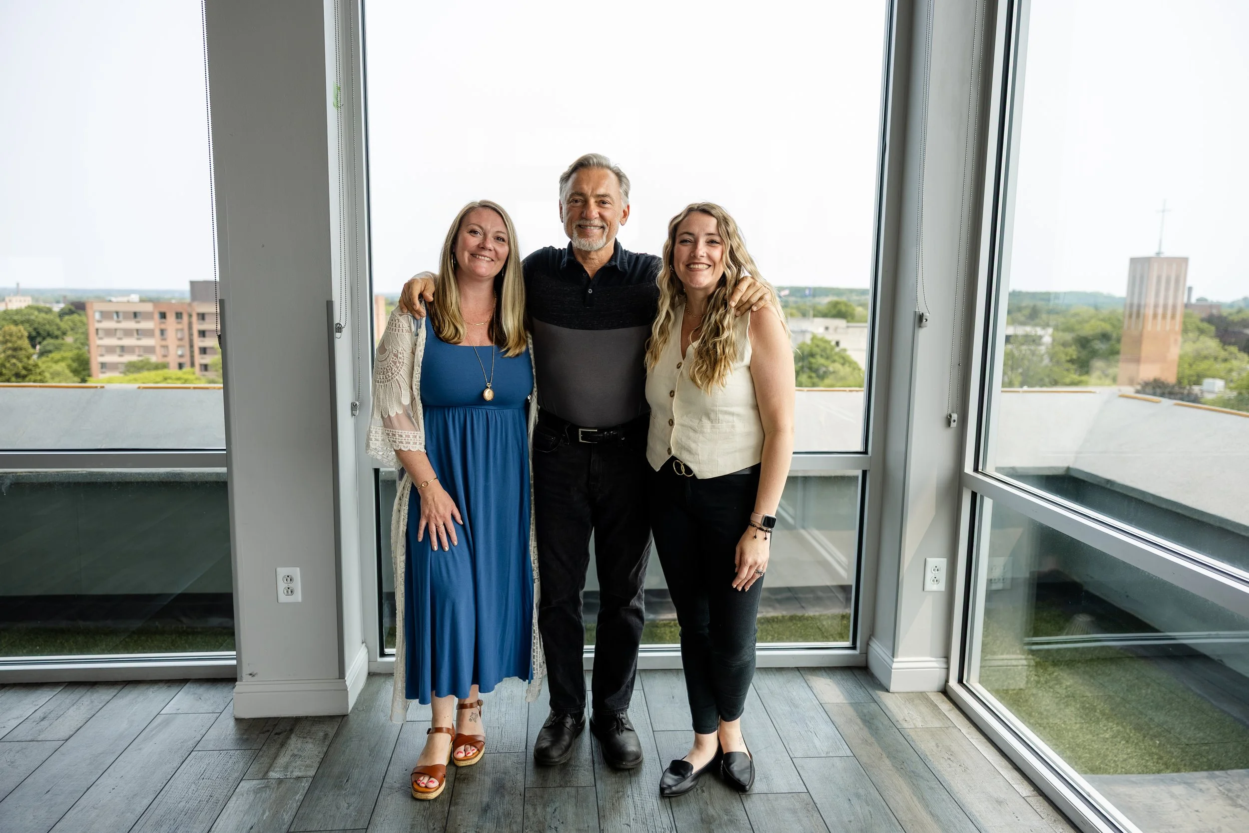Three people stand close together inside a room with large windows, showing a cityscape and greenery outside. They are smiling and have their arms around each other.