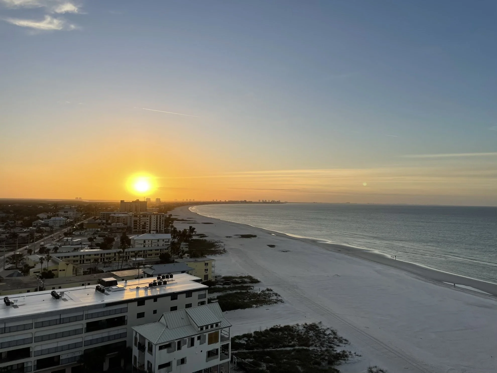 Sunset over a beach with calm ocean waves, sandy shore, and buildings along the coastline.