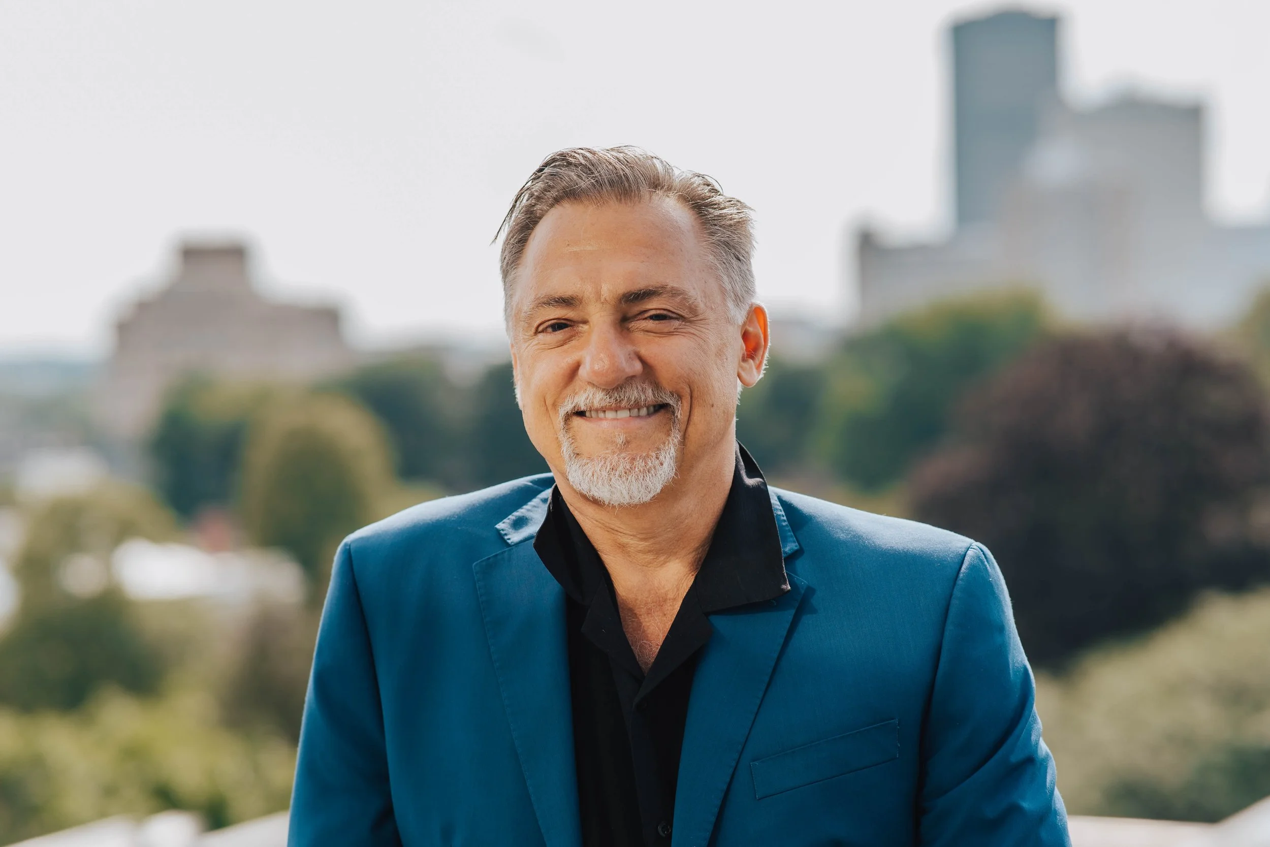 A middle-aged man with gray hair and a beard, wearing a blue blazer and a black shirt, smiling outdoors with a cityscape and trees in the background.