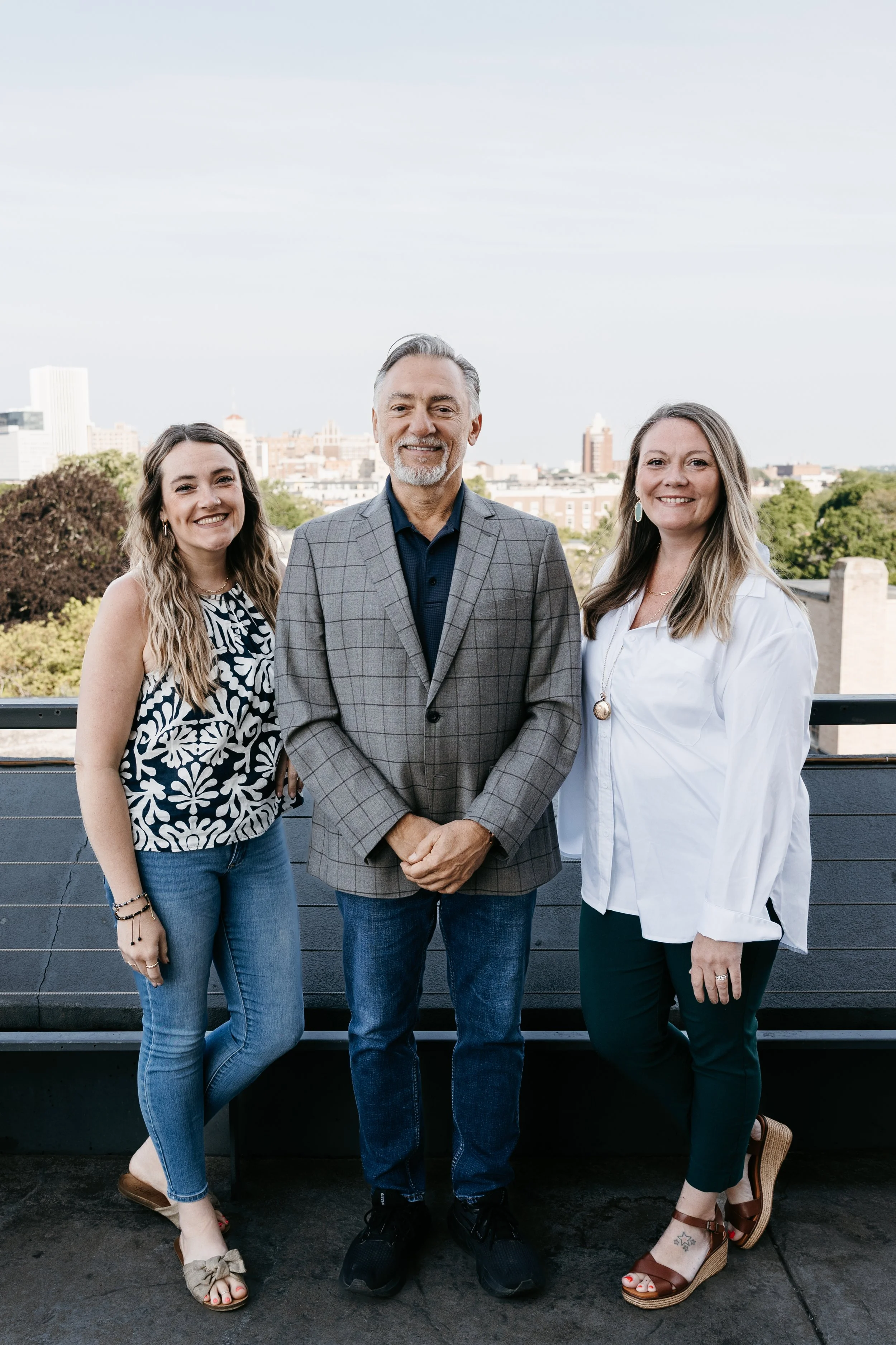 Three people standing outdoors on a rooftop with a city skyline in the background, smiling at the camera.