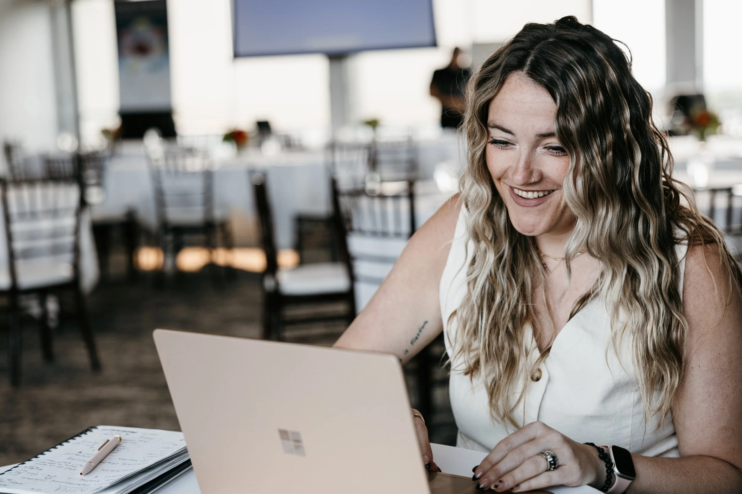 A woman with curly hair and a nose piercing smiling while working on a laptop in a bright restaurant or cafe with tables and chairs in the background.