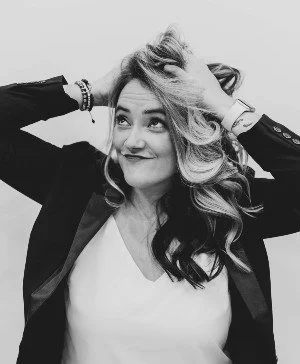 Black and white photo of a woman smiling with her hands in her hair, wearing a blazer and a white top.