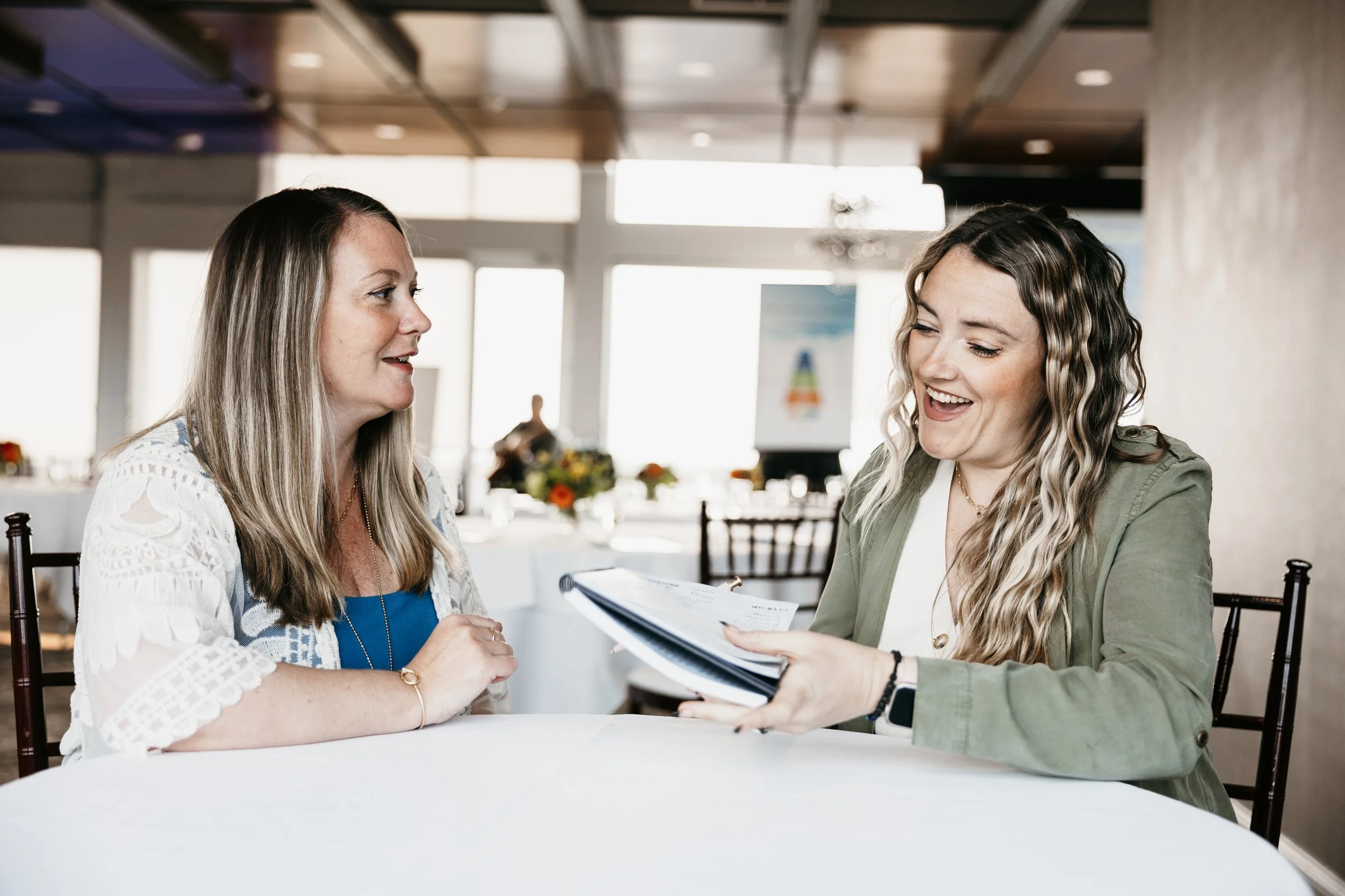 Two women sitting at a table, smiling, engaged in conversation, with a blurred background of a bright indoor setting
