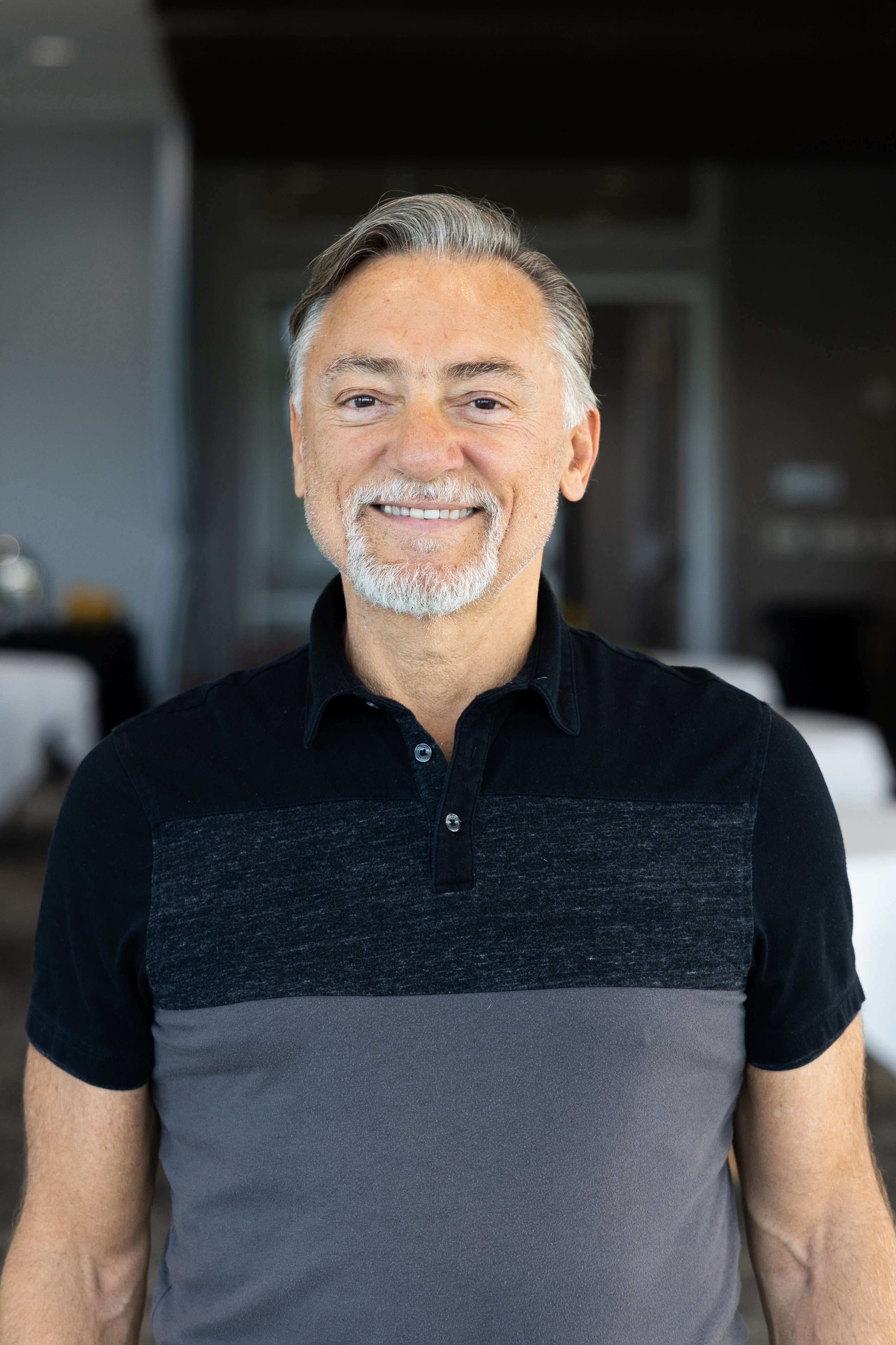 A smiling middle-aged man with gray hair and beard wearing a blue blazer and black shirt, standing outdoors with a city skyline and greenery in the background.