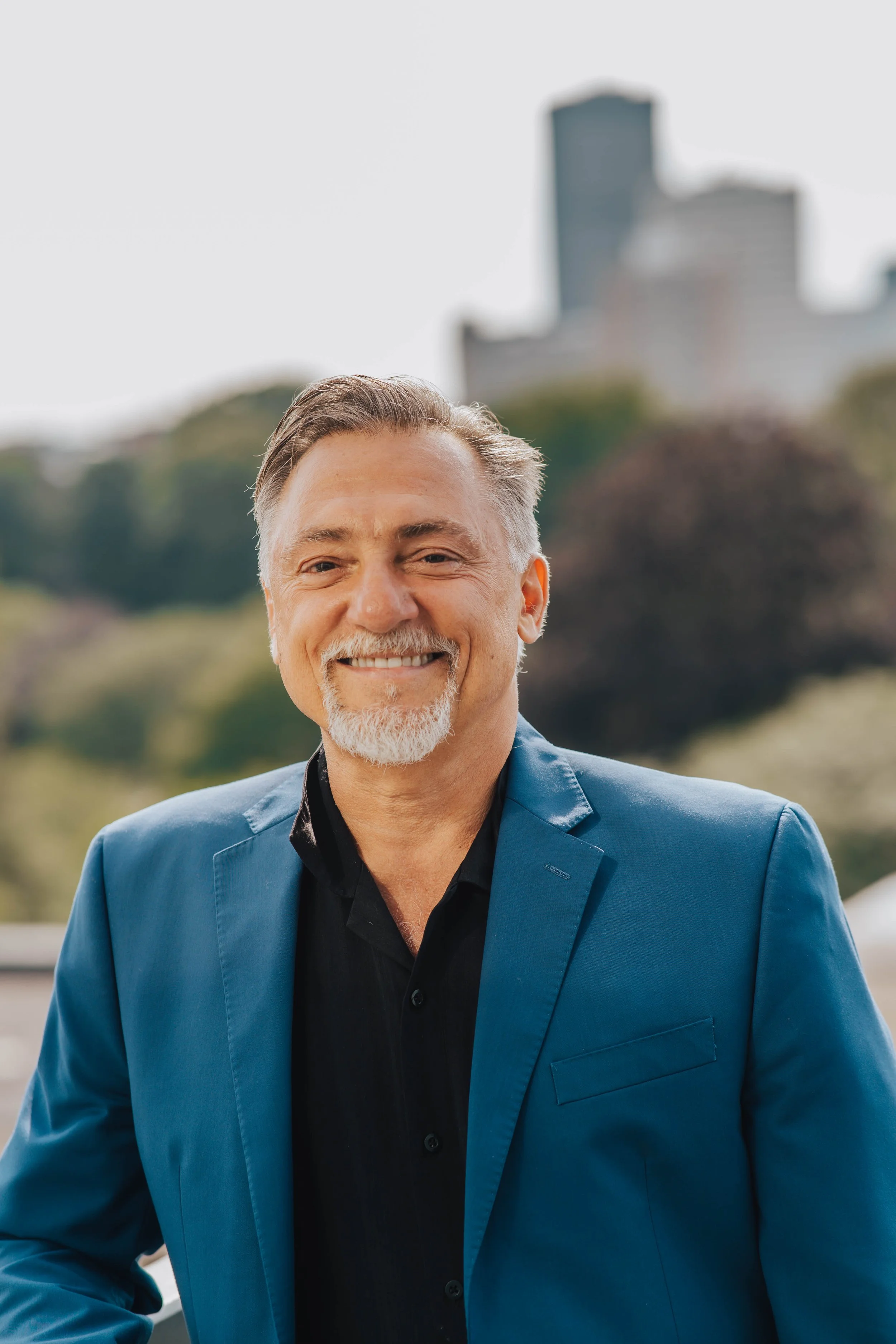 A smiling middle-aged man with gray hair and a beard, wearing a blue blazer over a black shirt, standing outdoors with a cityscape and trees in the background.