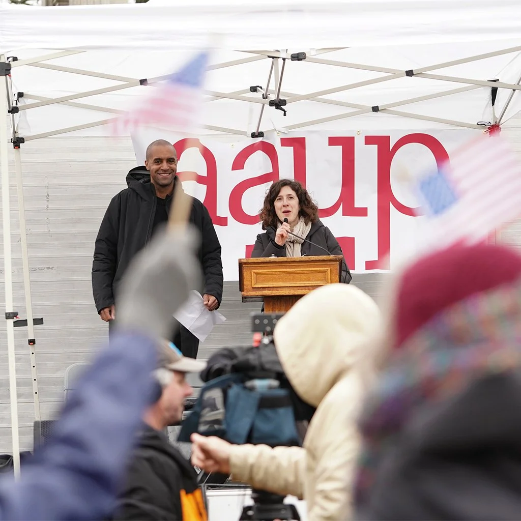 A woman speaking at a podium during a rally or outdoor event, with a man standing beside her and a crowd in front holding flags. There is a large banner behind them with partially visible red text.