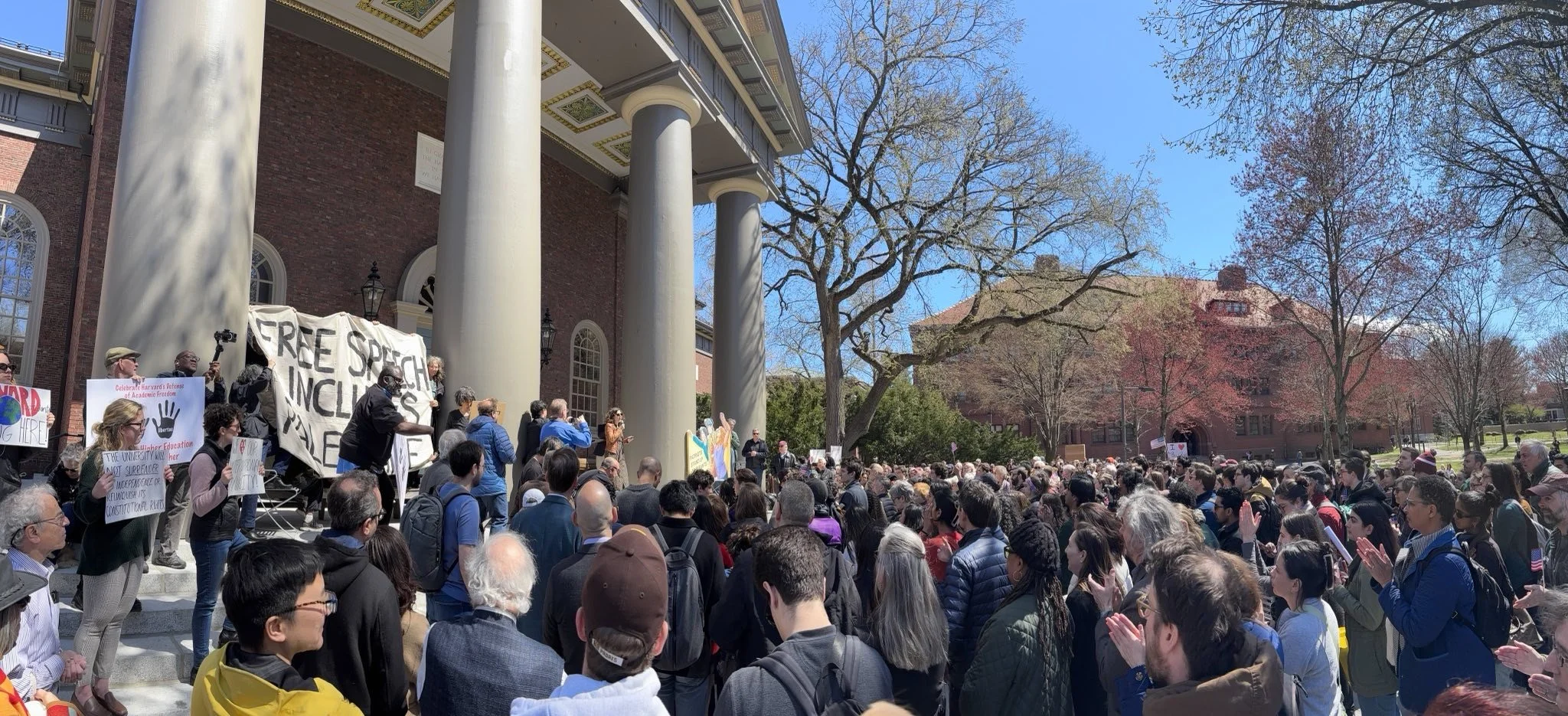 Crowd gathered outside a red brick building with large columns, holding signs and listening to a speaker at a rally on a sunny day with leafless trees.