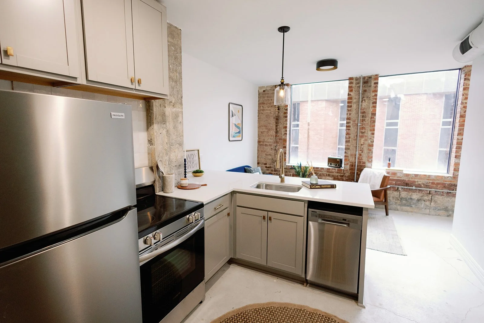 Modern kitchen with stainless steel appliances, white cabinetry, a large window, an island with a sink, and exposed brick walls in an urban apartment.