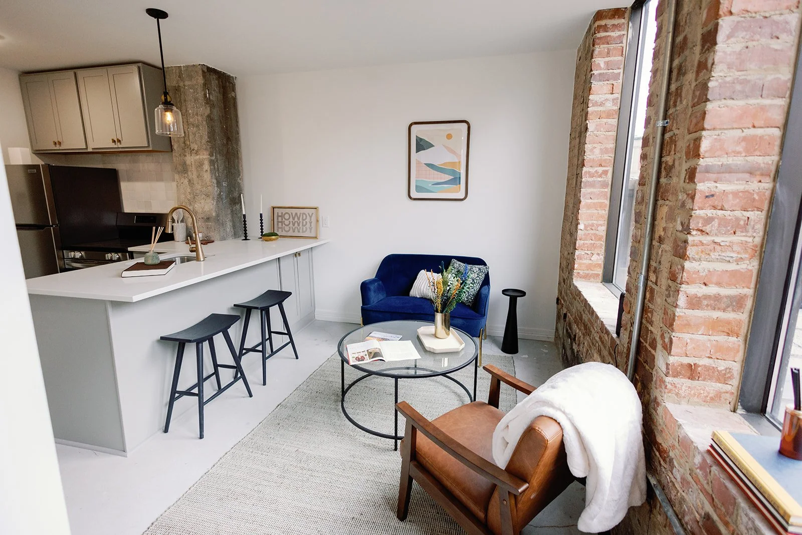 Modern apartment living room with exposed brick wall and large window, featuring a blue velvet armchair, wooden armchair with a white blanket, round glass coffee table with magazines and a flower vase, kitchen bar with two stools, and decorative art