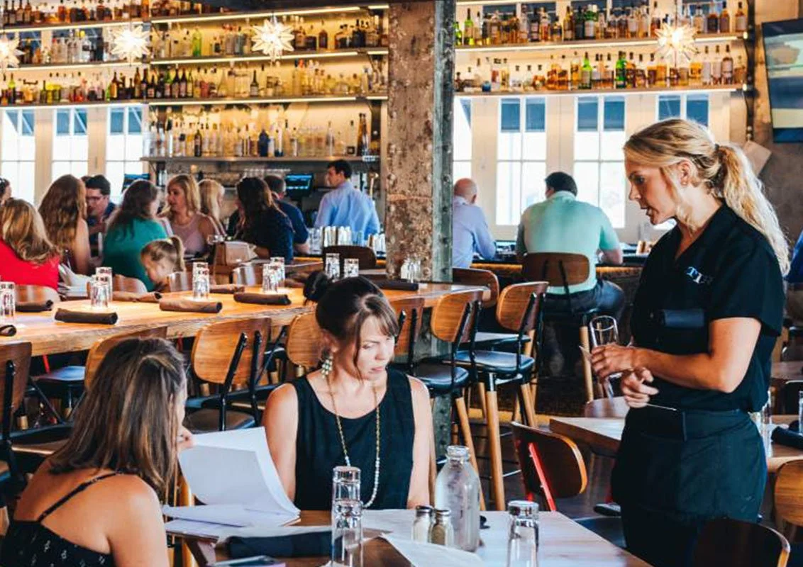 A busy restaurant with multiple people dining and a bartender or waitress taking an order, with shelves of liquor bottles and bar equipment in the background.