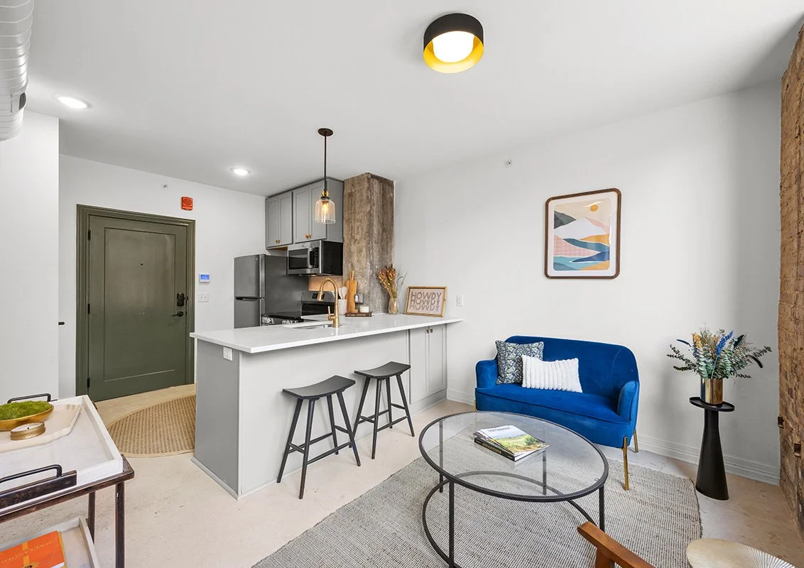 Modern living room with a blue velvet sofa, glass coffee table, and wall art, adjacent to a kitchen with gray cabinets, stainless steel appliances, and a white countertop island with two black stools.