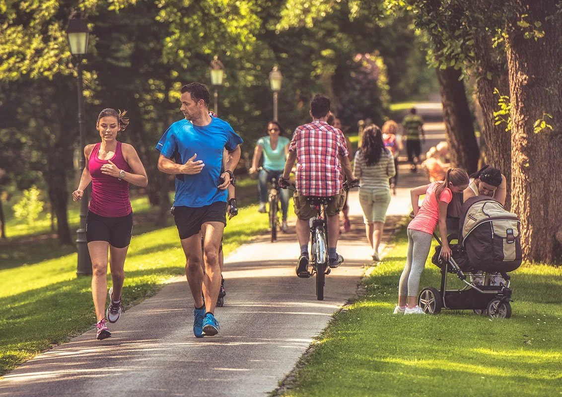 People enjoying outdoor activities in a park on a sunny day, including running, cycling, and a woman with a child in a stroller.
