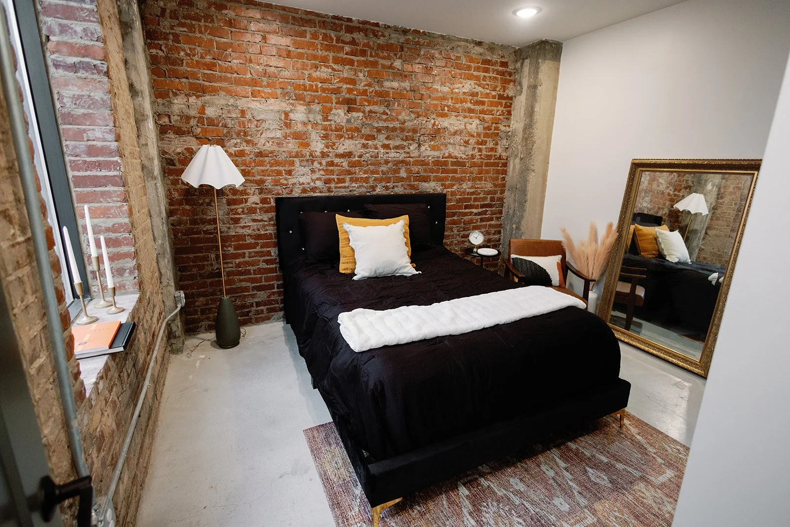 A bedroom with an exposed brick wall behind a black bed, white and yellow pillows, a white blanket, a standing lamp, a small side table, a large gold-framed mirror, a chair with orange and black pillows, a vase with dried plants, and a window with a