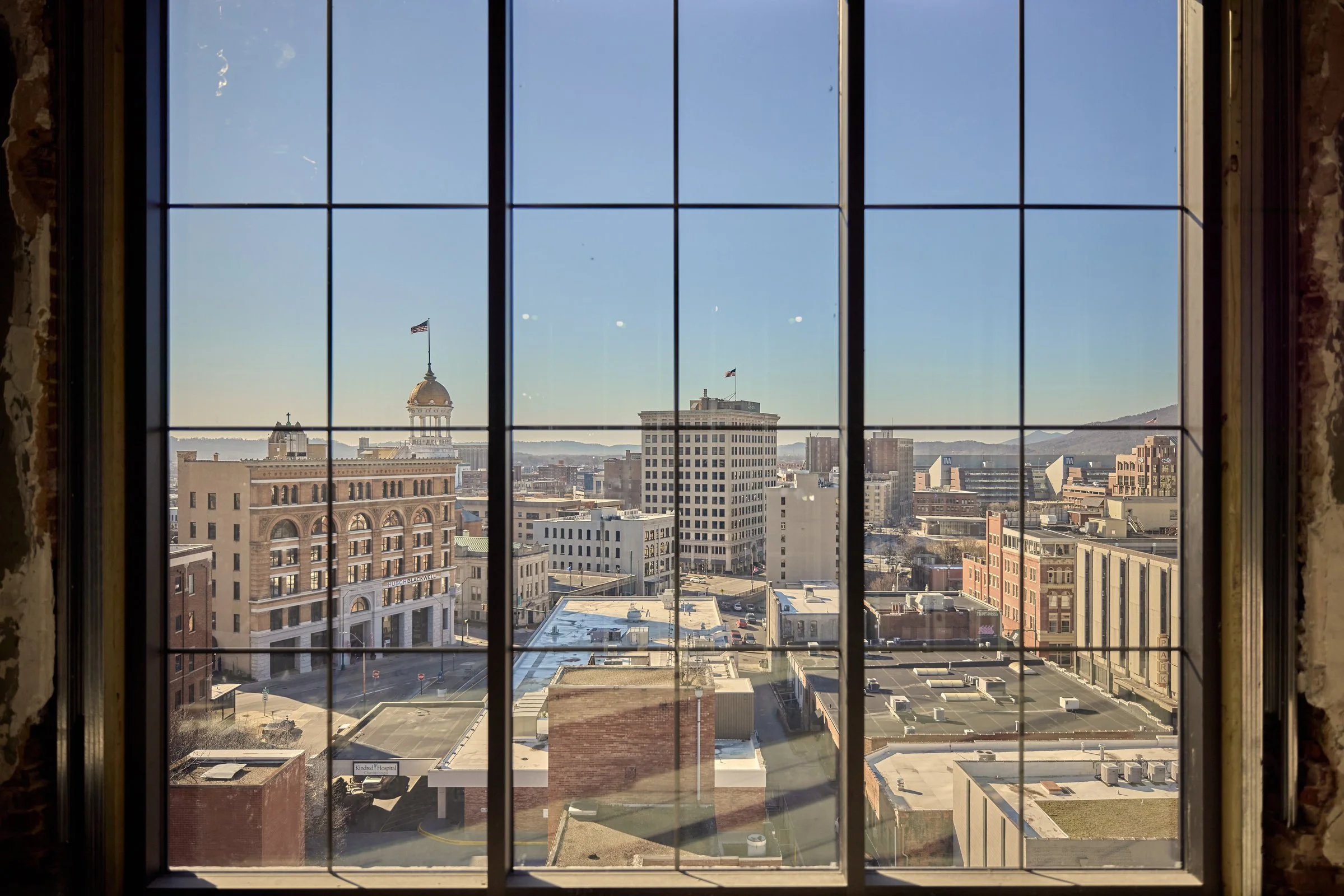 View of a city skyline through a window with grid panes, showcasing historic buildings with American flags, modern structures, and hills in the background, under a clear blue sky.
