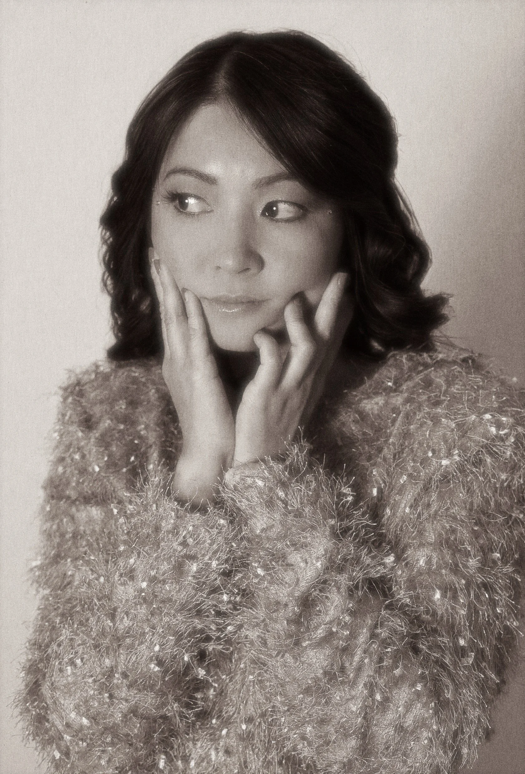 A woman with dark, shoulder-length hair styled in waves, wearing a textured, sparkly top, and resting her hands on her face.