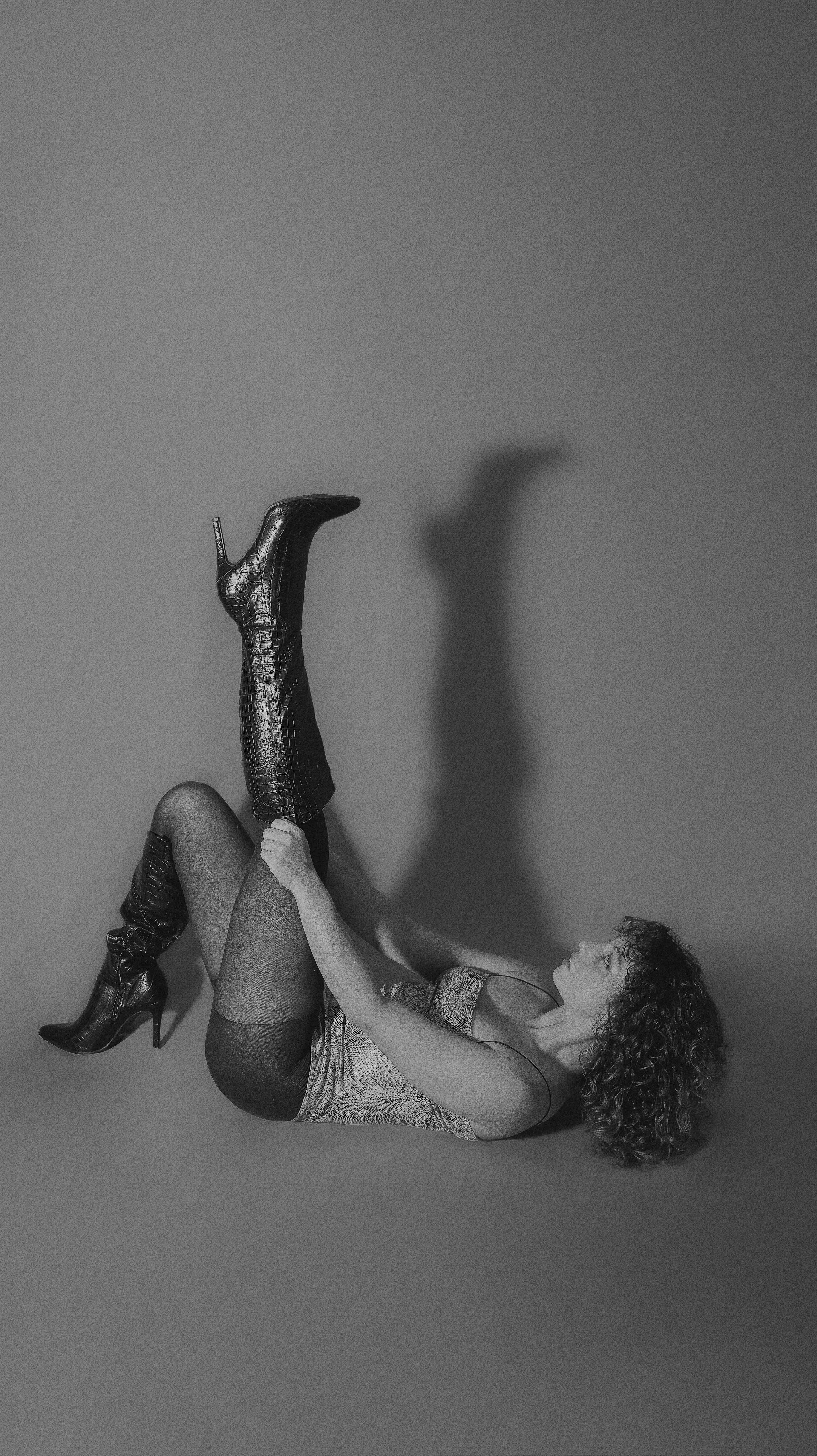 A woman lying on the ground in black and white, holding one leg up vertically while wearing high-heeled boots and fishnet stockings, with her head tilted back and curly hair.
