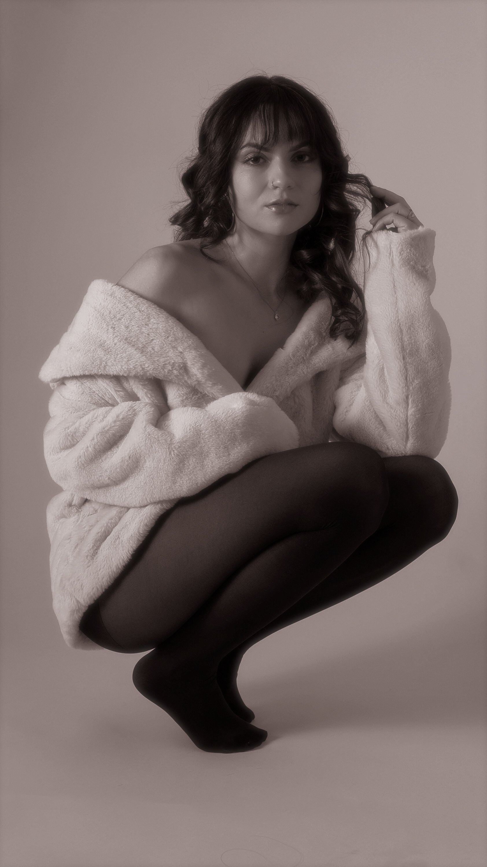 A woman with dark wavy hair wearing a white fuzzy coat and black tights, crouching against a plain background.