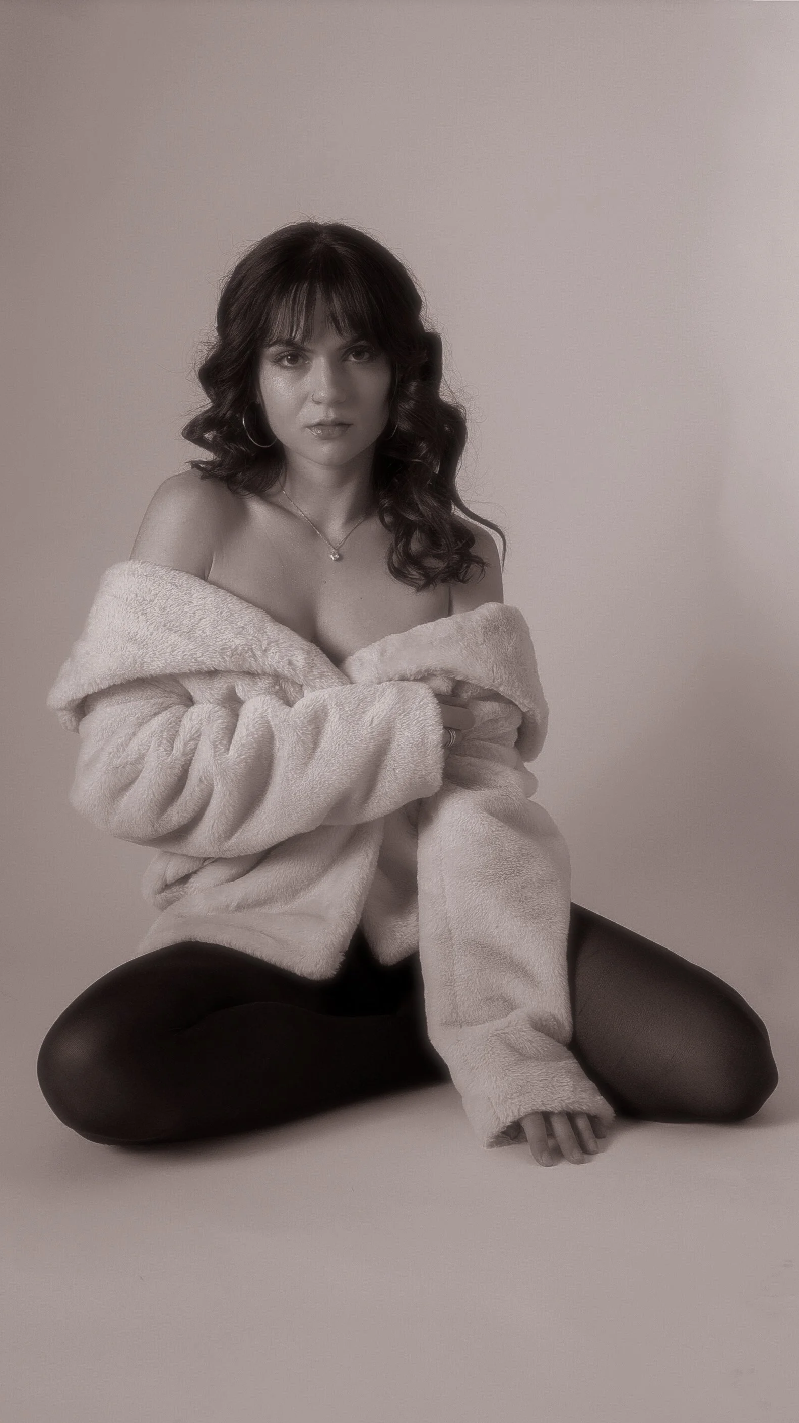 A woman with dark curly hair wearing a plush off-shoulder coat, sitting cross-legged on the floor against a plain background.