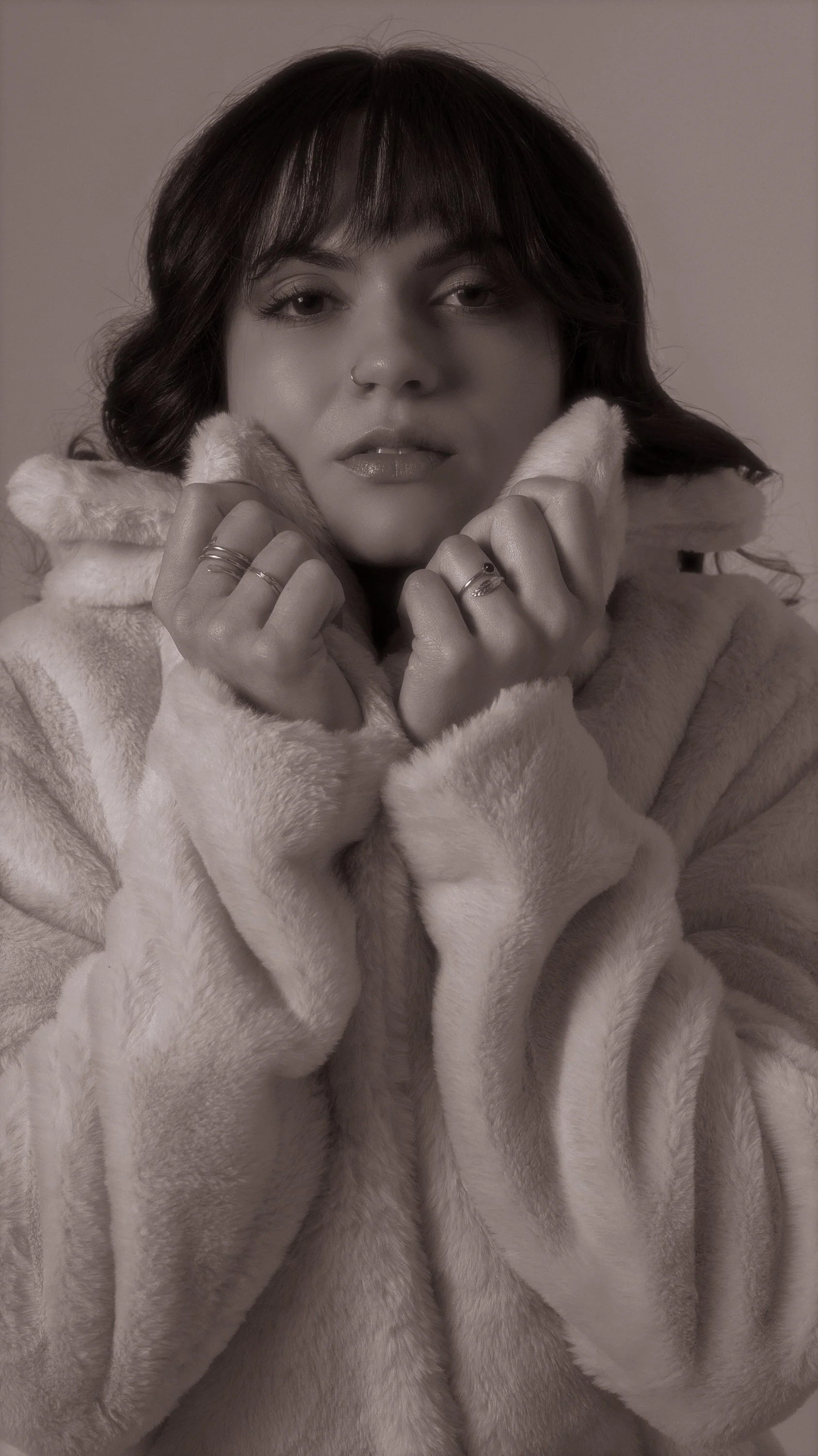 A woman with dark hair and bangs wearing a light-colored faux fur coat, holding its collar with both hands, looking at the camera.