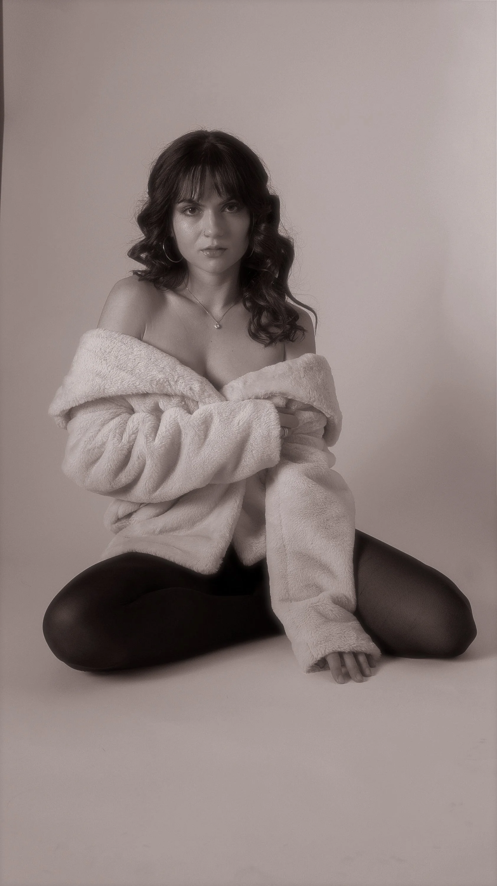 A woman with dark curly hair sitting on the floor with her legs crossed, wearing black tights and a fluffy light-colored coat, against a plain light background.