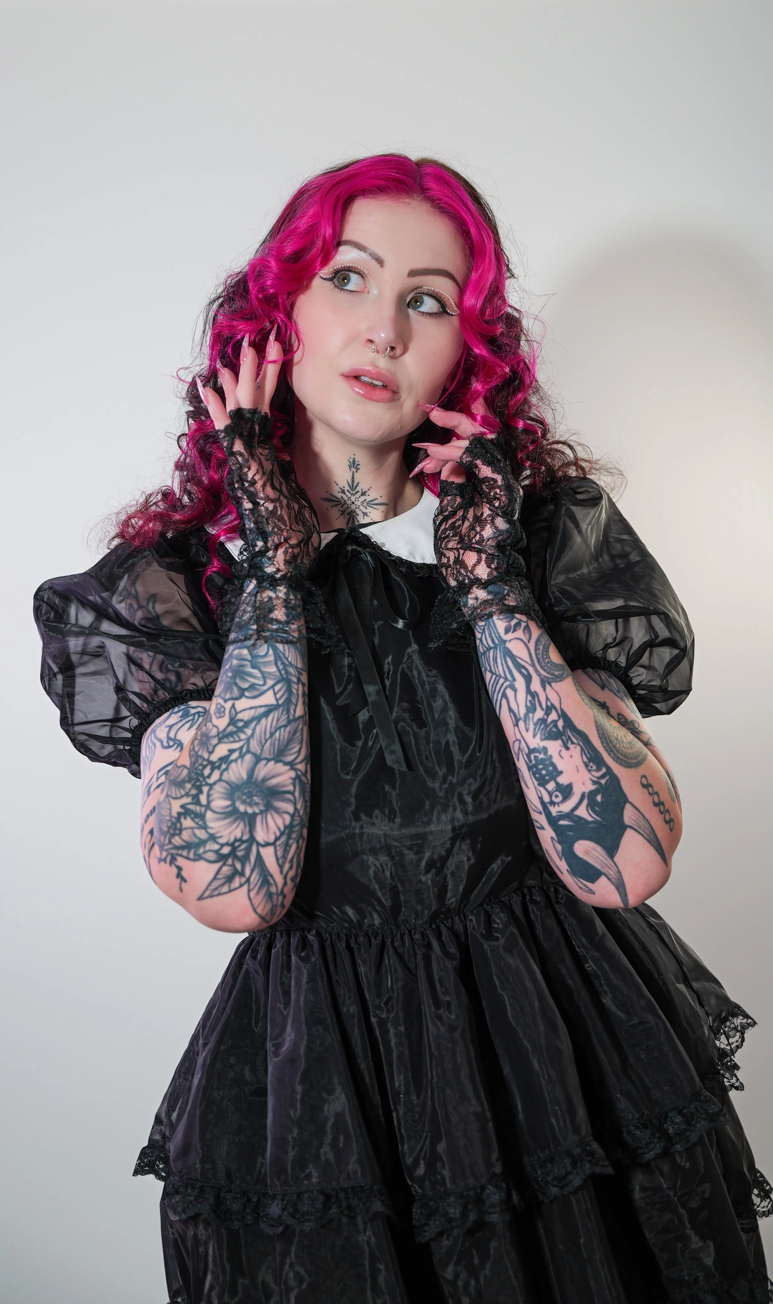 A woman with pink and black curly hair, tattoos on her arms, and wearing lace gloves, standing against a plain background, dressed in a black gothic dress with puffed sleeves and lace details.