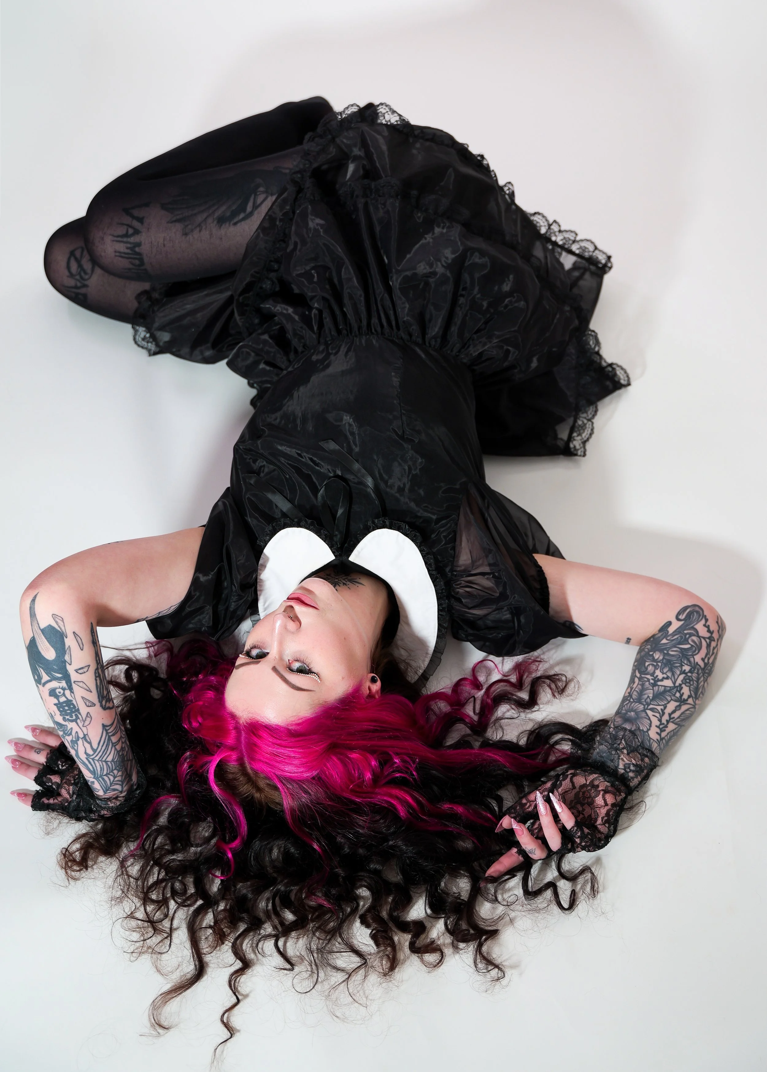 A woman with pink and black curly hair lying on a white surface, wearing a black dress with lace gloves and tattoos on her arms.