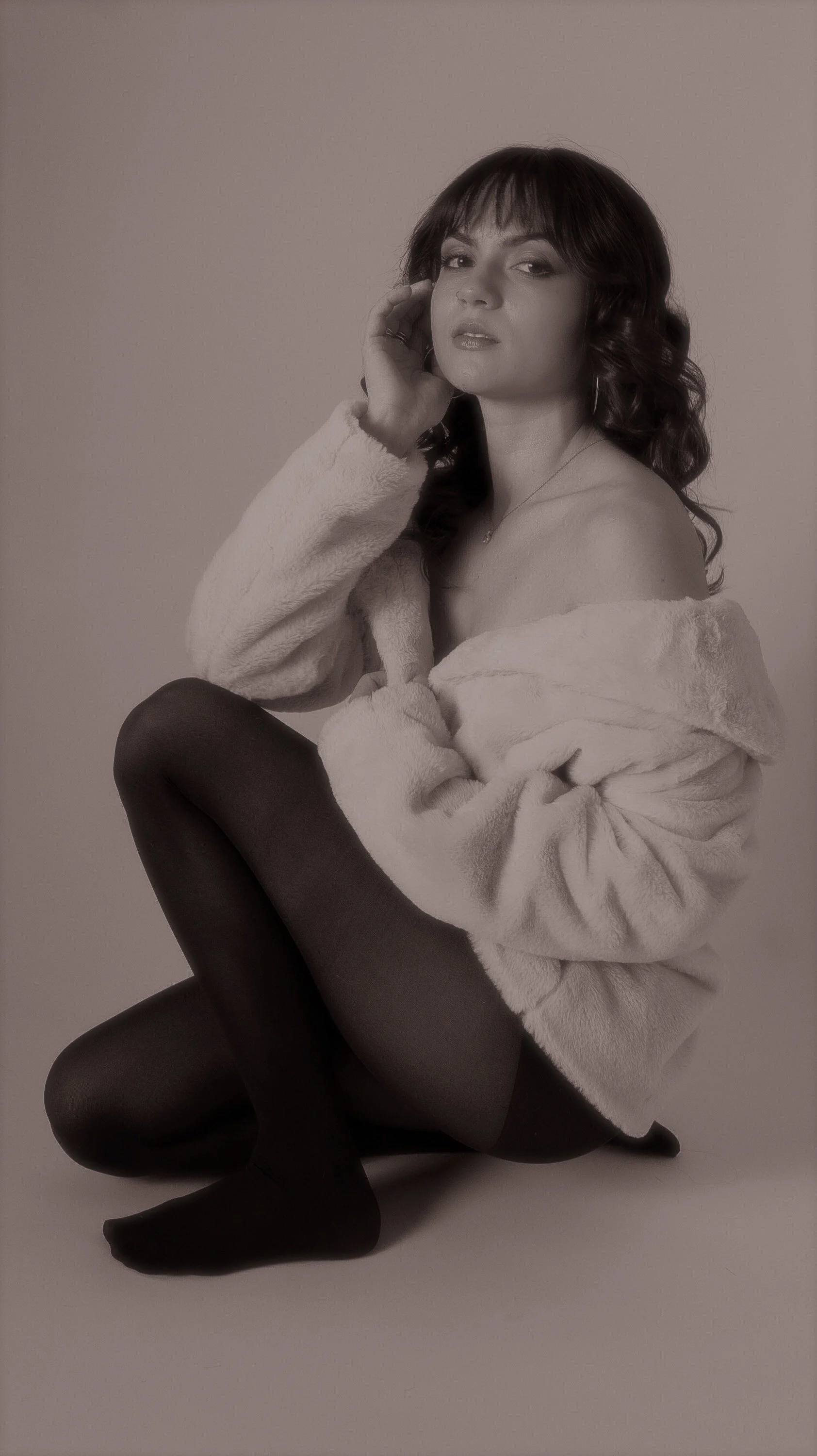 A woman with dark, curly hair sitting on the floor with one knee up, wearing dark tights and a plush, off-the-shoulder coat, looking at the camera.