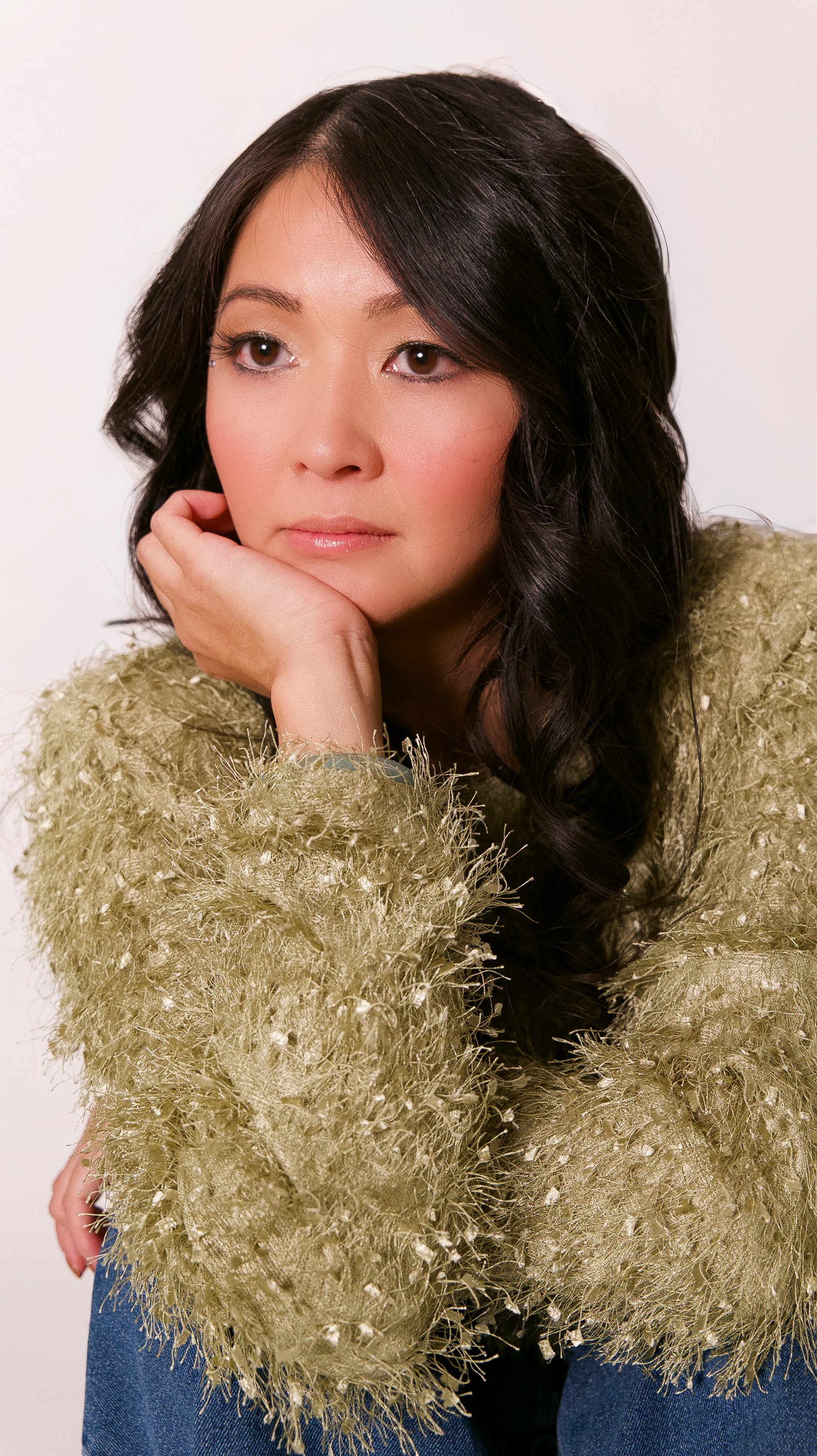 A woman with long dark hair and light skin, resting her chin on her hand, wearing a textured gold-colored sweater and blue jeans, posing against a white background.