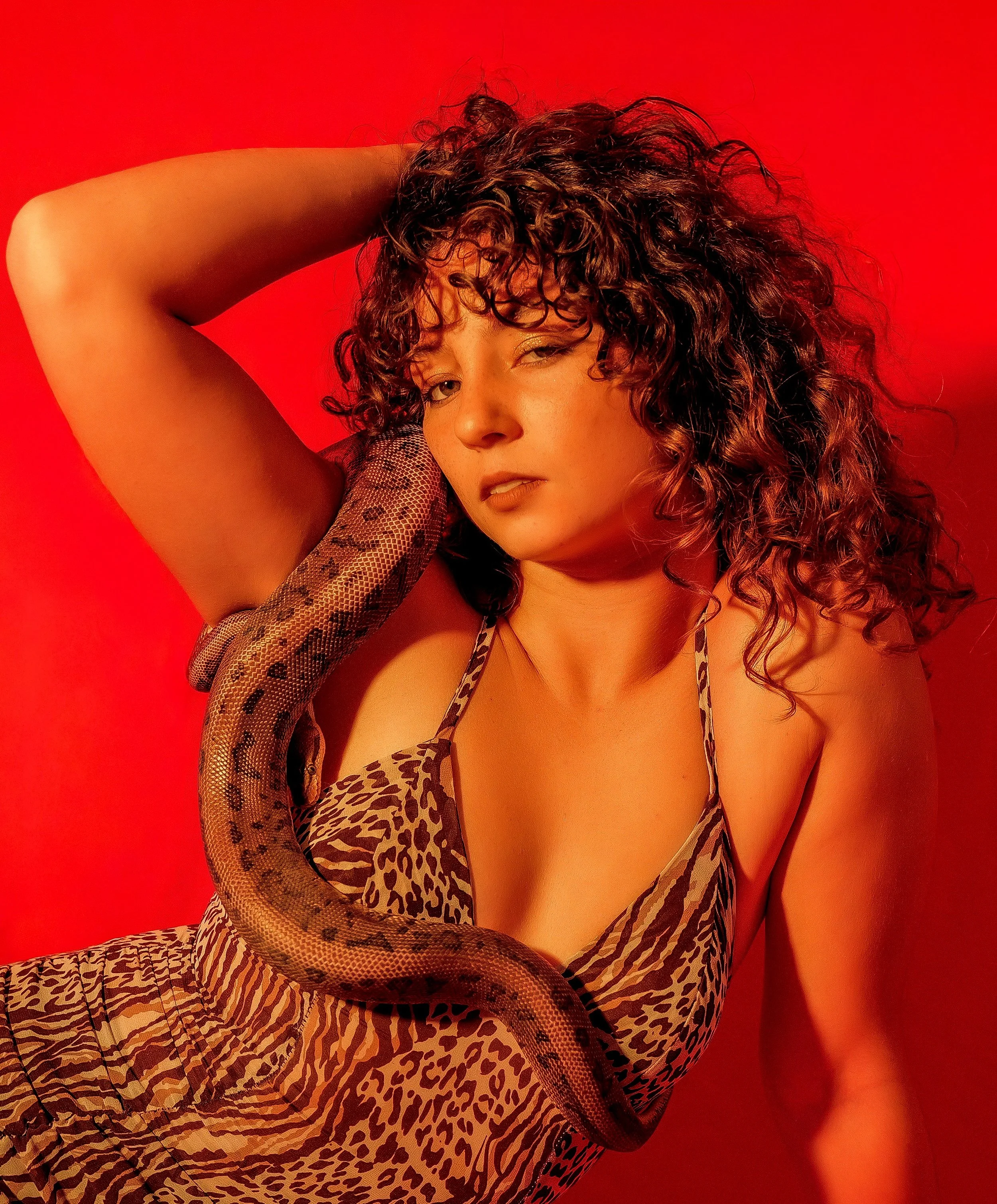 A woman with curly hair in a leopard print dress holding a snake on her arm, standing against a red background.