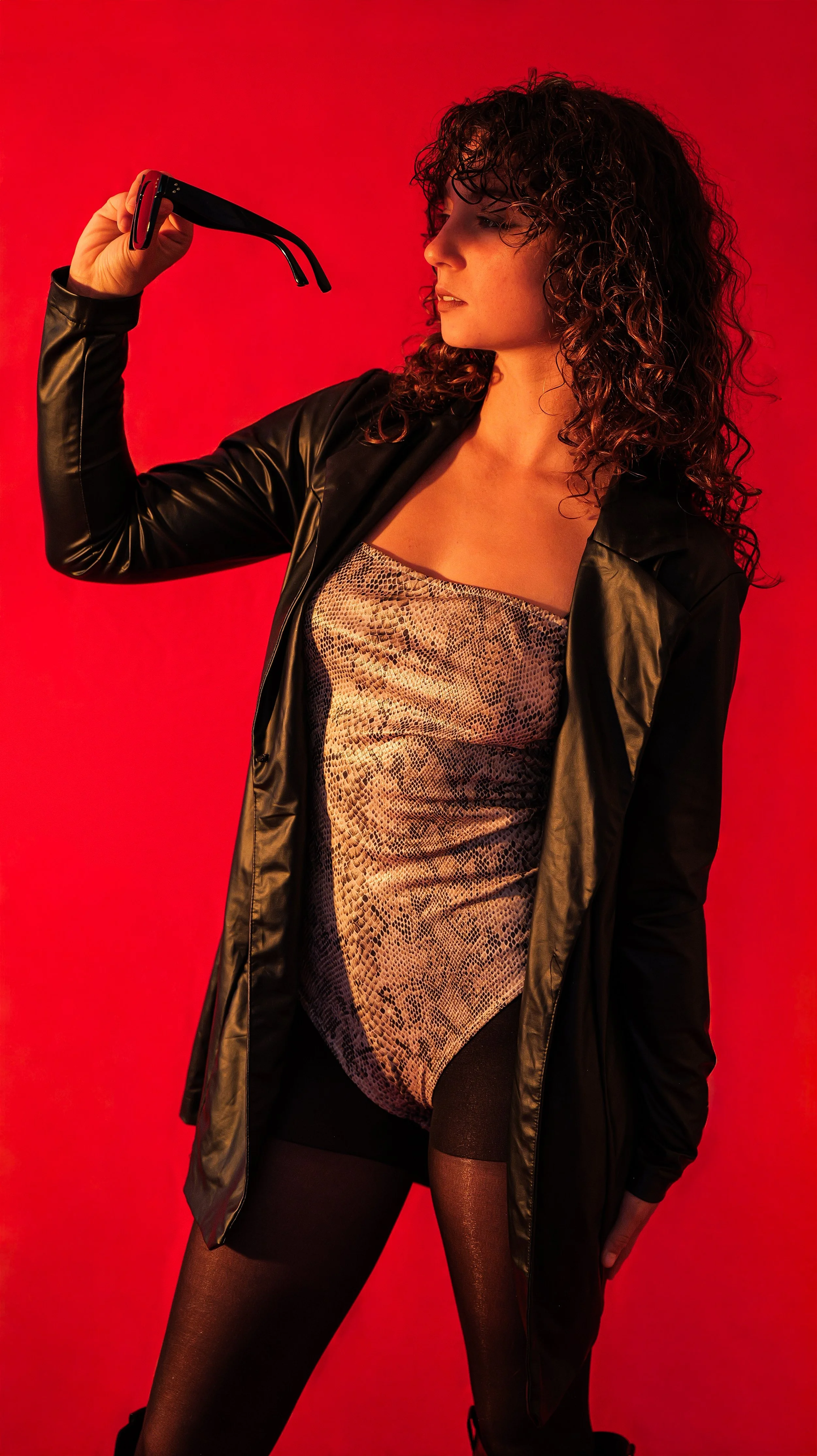 A woman with curly hair in a black jacket and shiny snake-patterned dress takes a selfie against a red background.