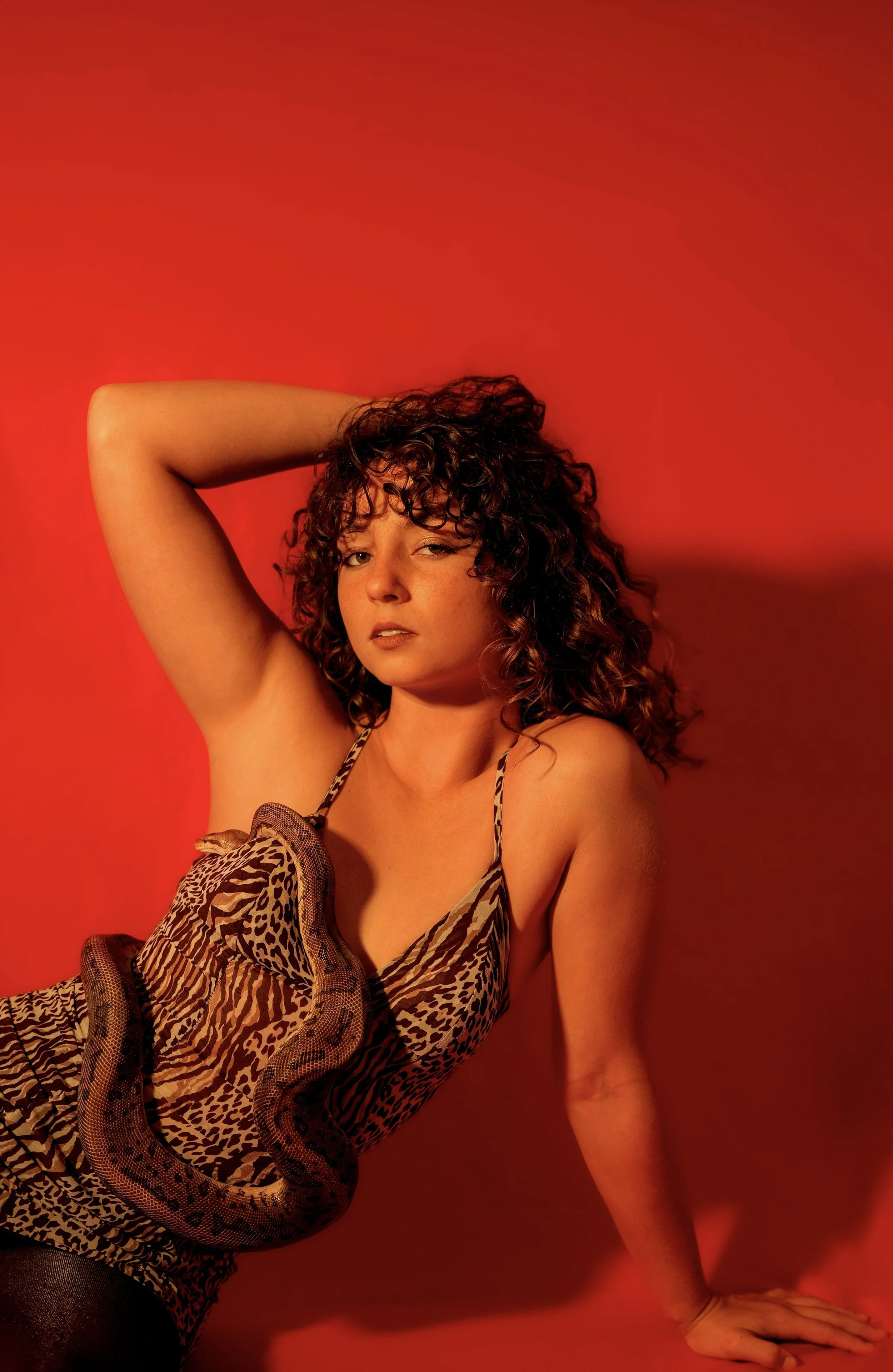 A woman with curly hair wearing a leopard print outfit, lying against a red background with a snake draped across her torso.