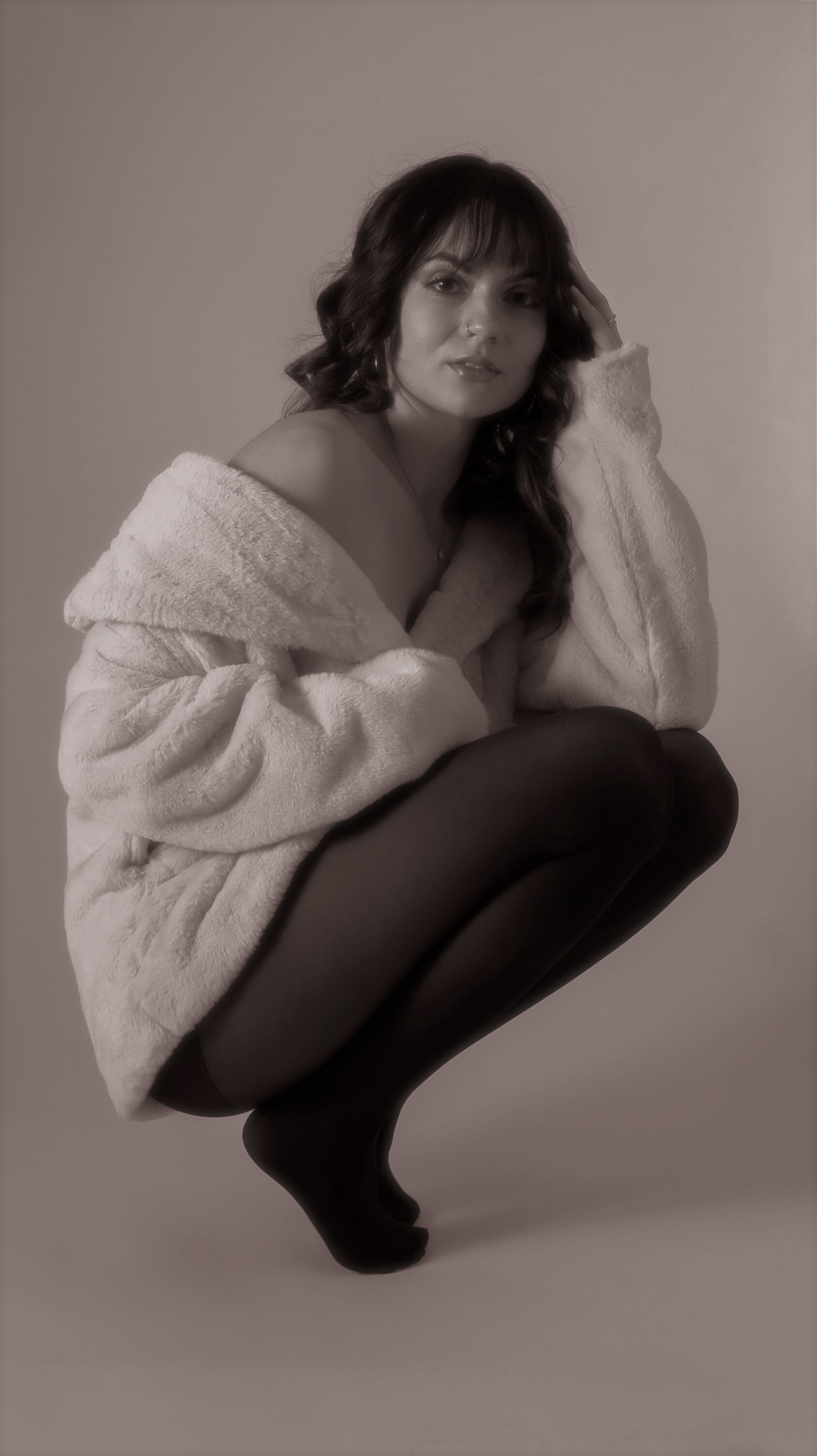 A woman with dark wavy hair, wearing a plush off-shoulder robe and black tights, squatting against a plain wall.