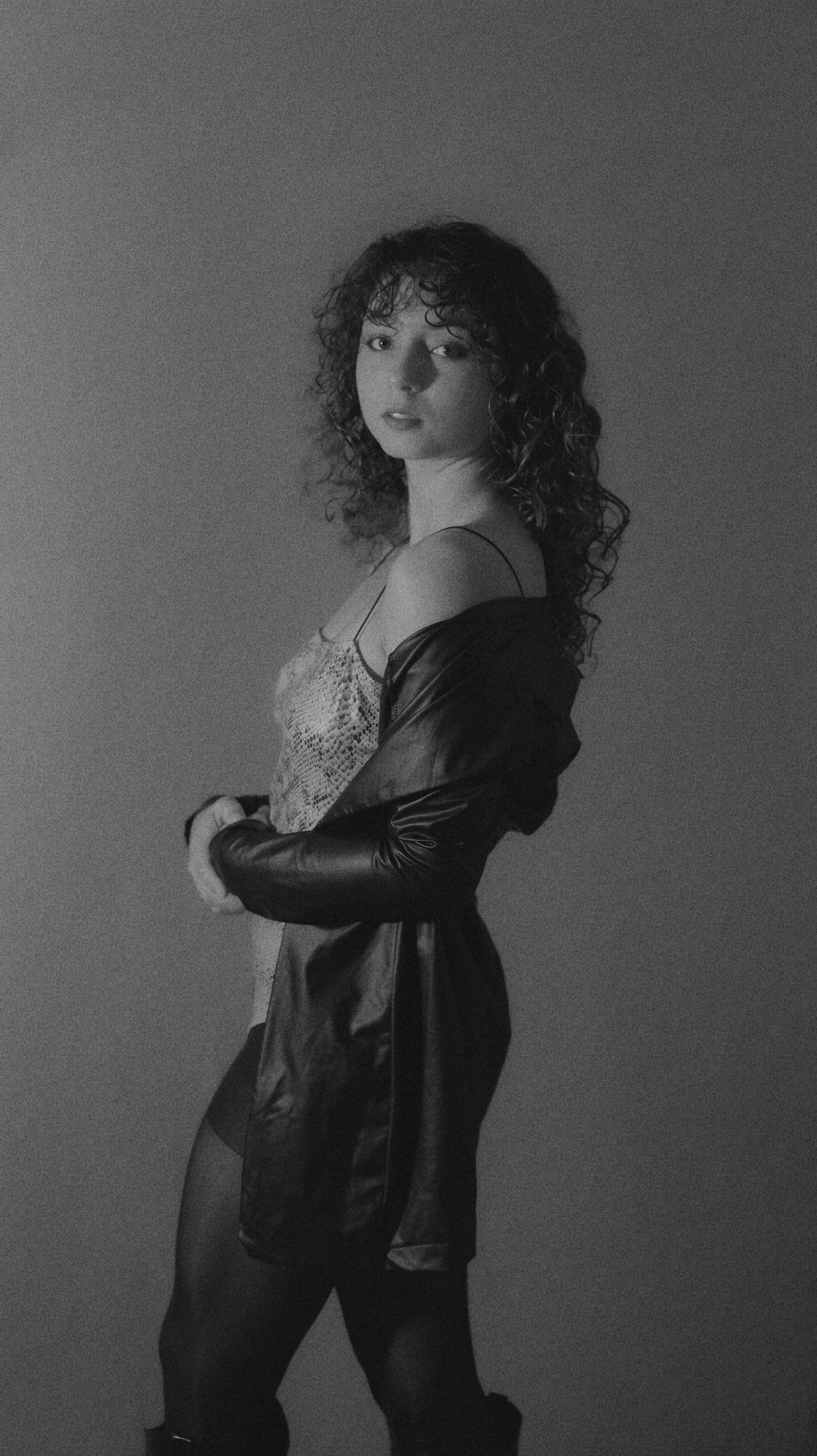 Black and white photo of a woman with curly hair wearing a snake-skin top, a black jacket hanging off her shoulders, and black tights, standing against a plain background. She is looking at the camera with a serious expression.