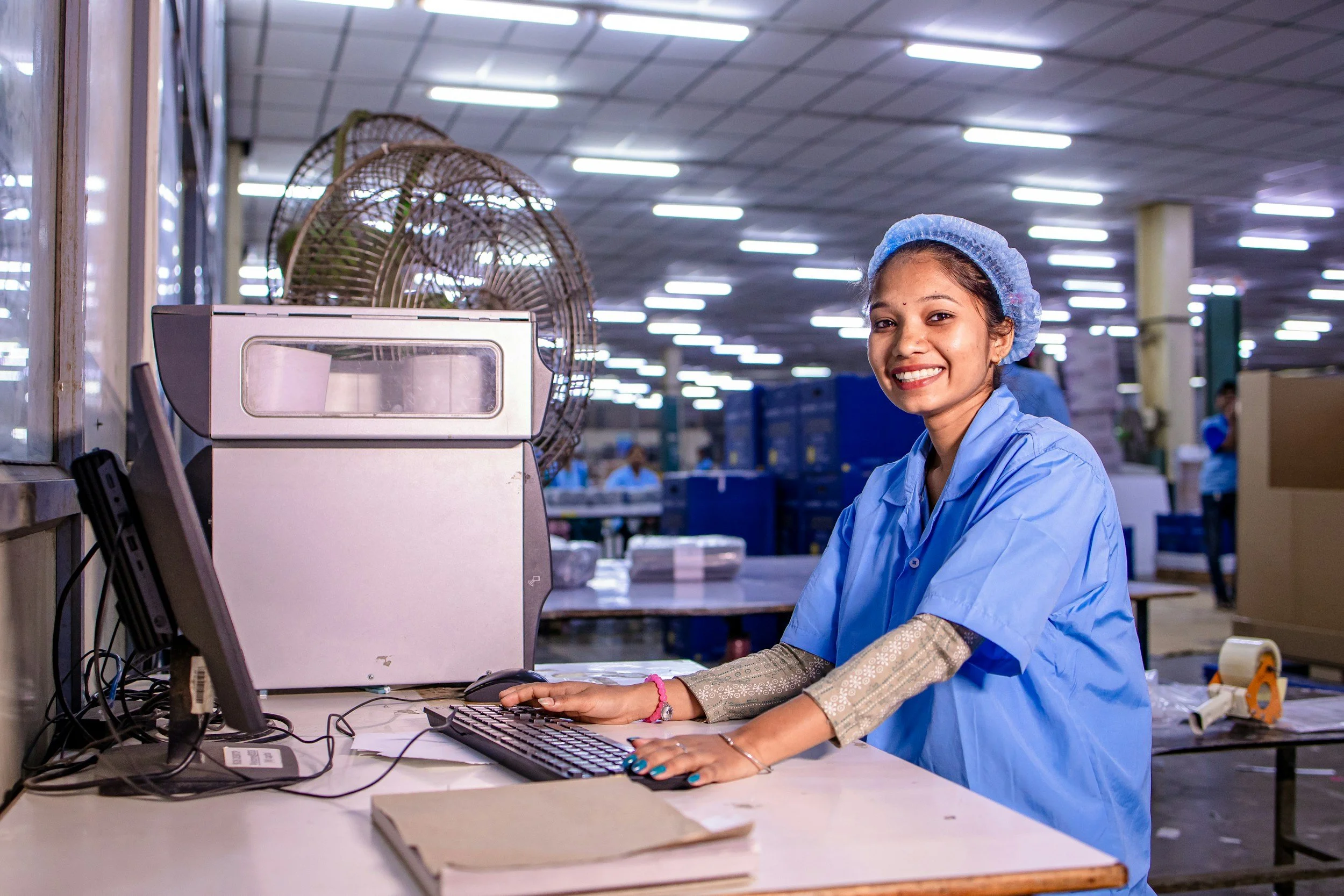A smiling woman in a blue protective gown and hairnet working at a desk with a keyboard and monitor in a factory or warehouse setting.