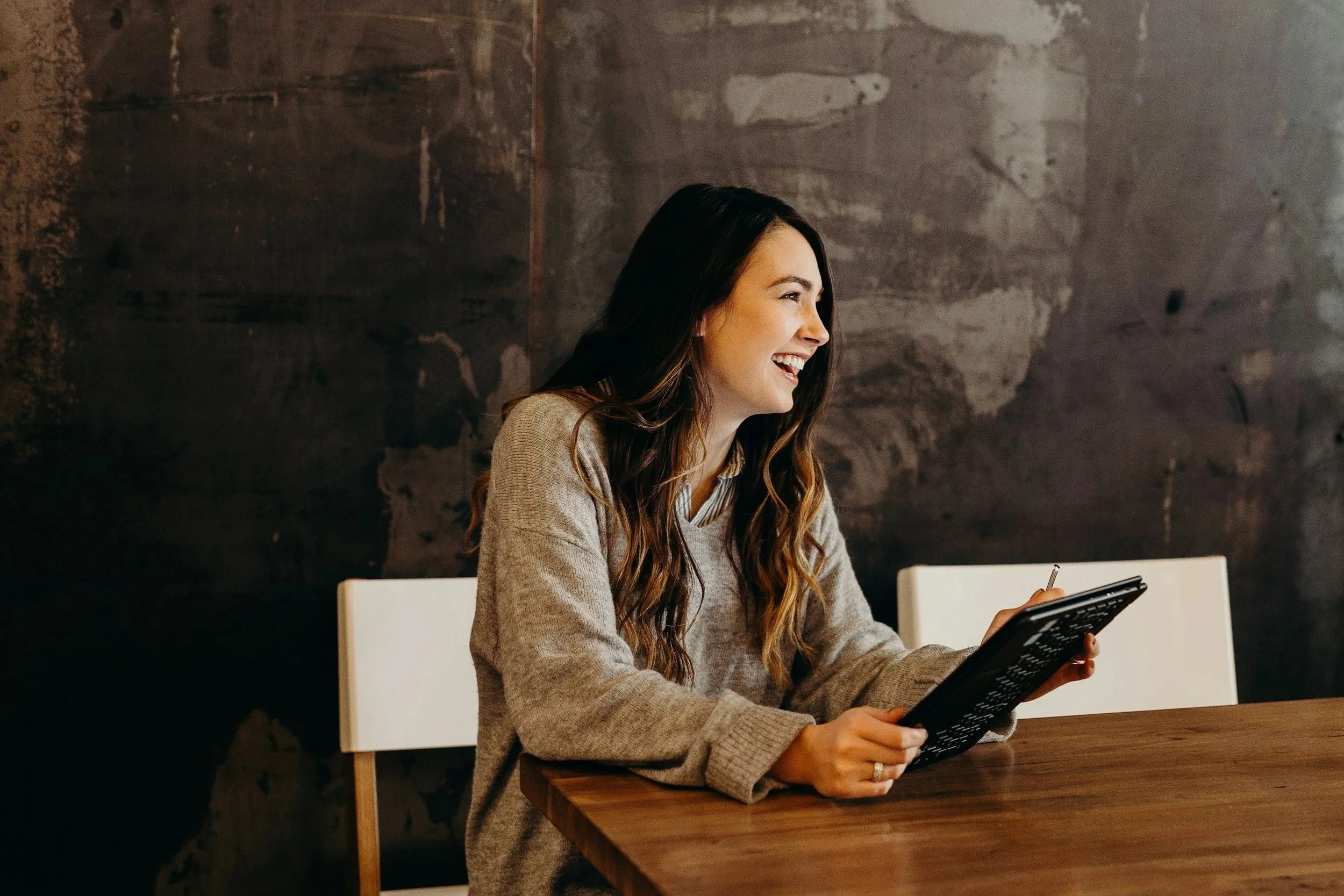 A woman with long wavy hair, wearing a beige sweater, is sitting at a wooden table and smiling while looking at a tablet she is holding in her hands.