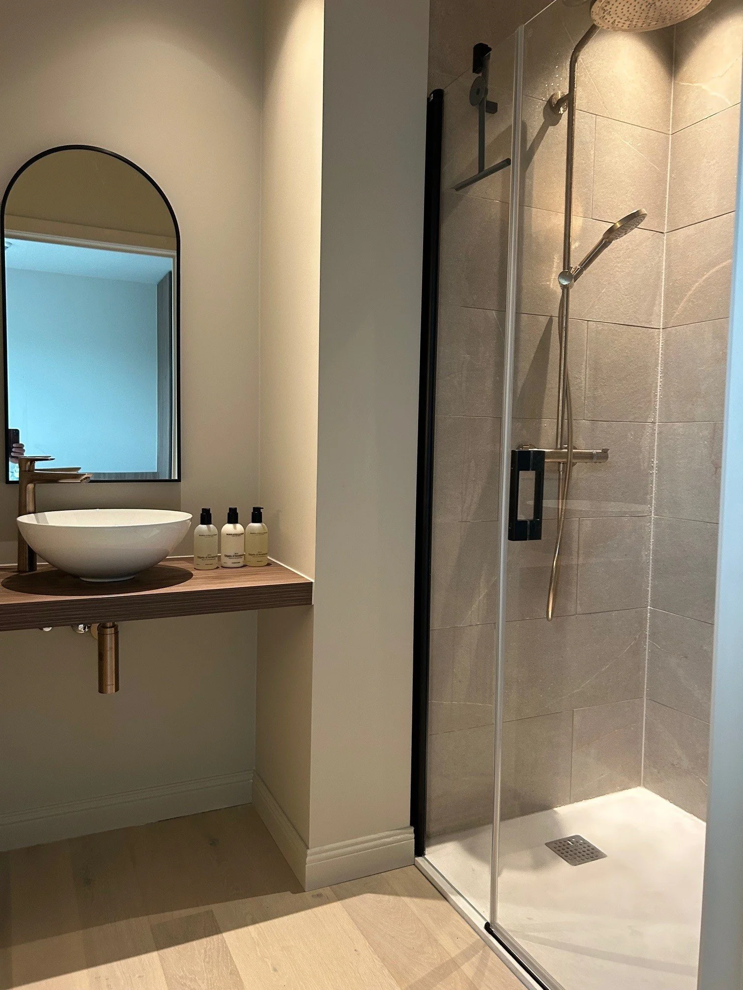 Modern bathroom with a vessel sink on a wooden floating vanity, three bottles of liquid soap or lotion, a mirror with an arched top, and a walk-in shower with gray tiles and a sliding glass door.