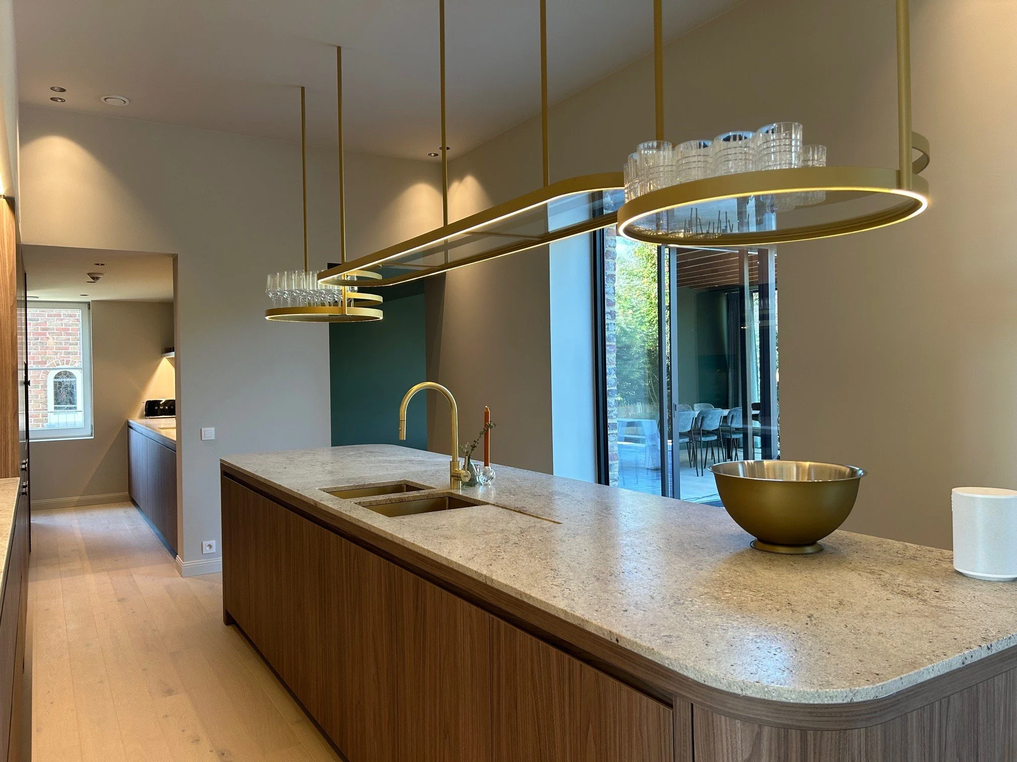 Modern kitchen with a large beige marble island, gold fixtures, and a view of a patio through sliding glass doors.