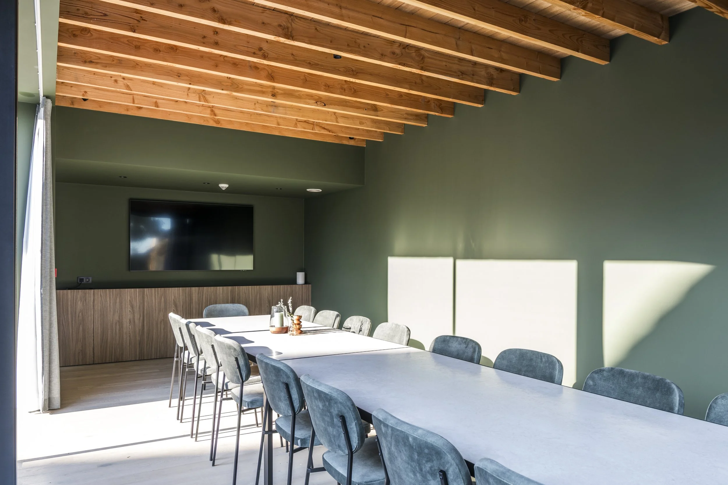 Modern conference room with a long beige table, green walls, wooden ceiling, electrical outlets, a large flat-screen TV, and sliding glass doors with curtains leading outside.