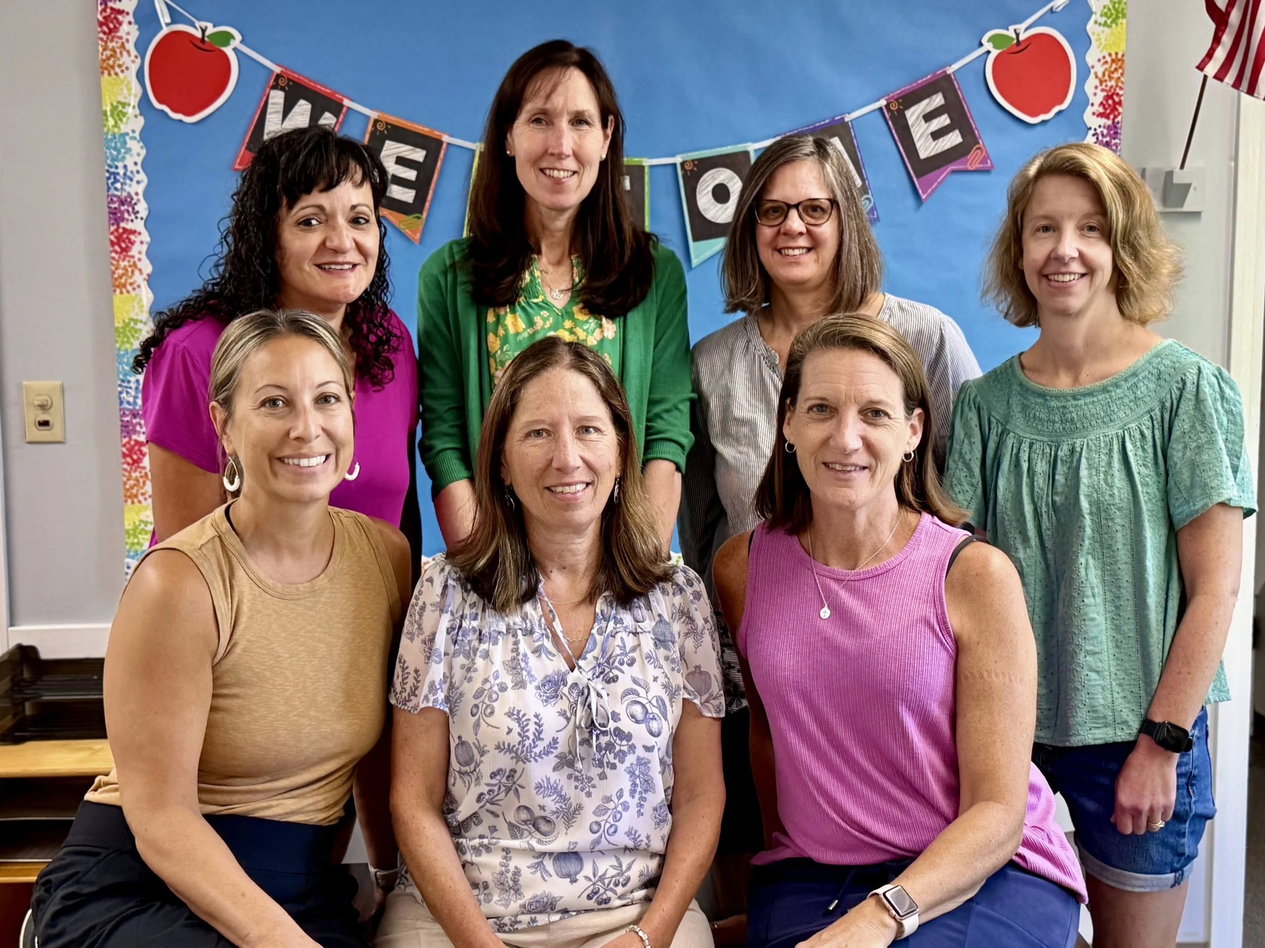 A group of eight female teachers posing together in a classroom with a colorful bulletin board, two of the women are seated and the rest are standing behind them.