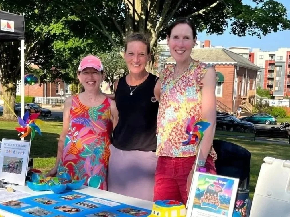 Three women standing behind a table at an outdoor event, smiling, with a tree and buildings in the background.