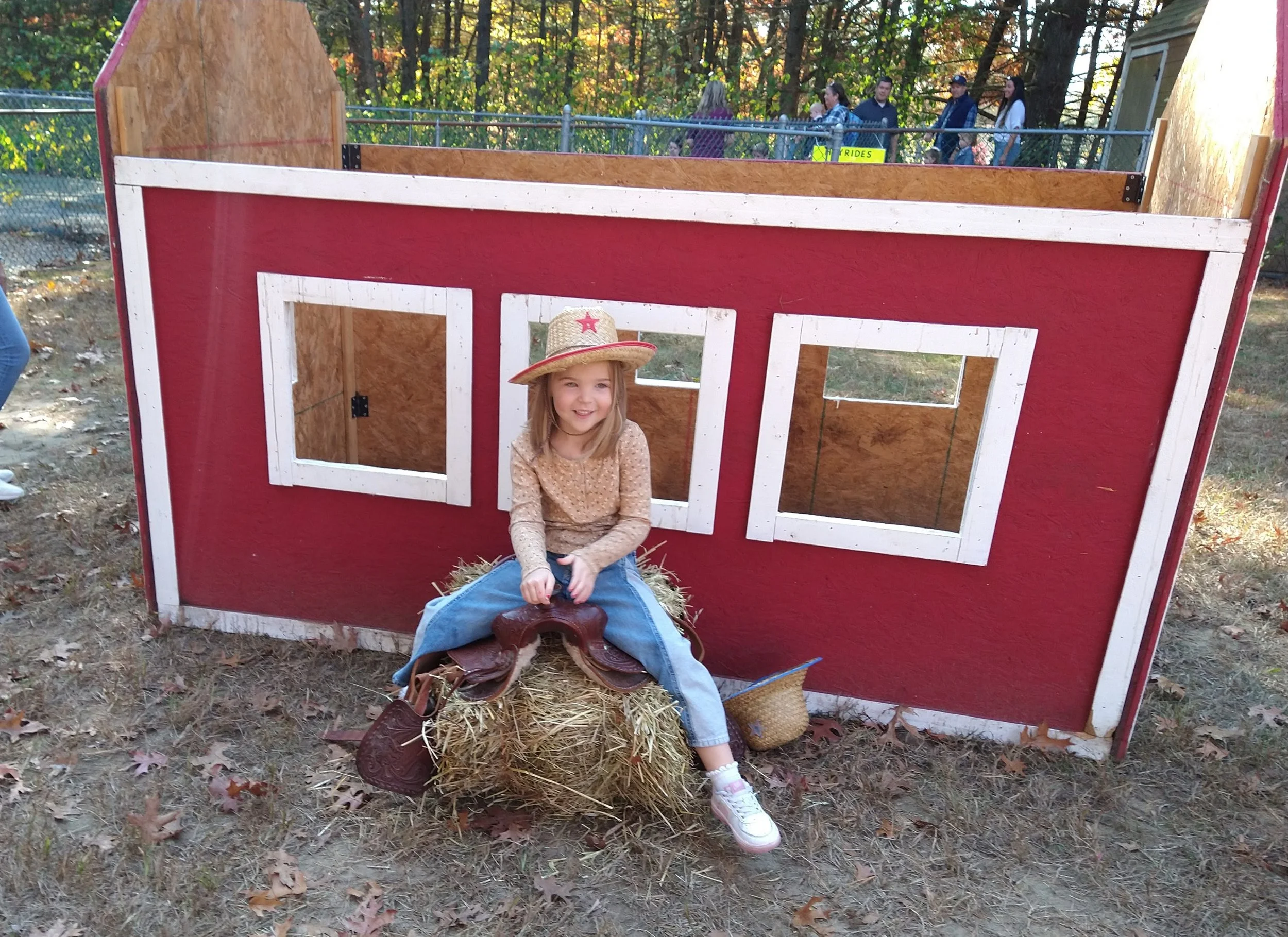 A young girl sitting inside a small red and white playhouse with a straw hat on her head, holding a cowboy hat and sitting on hay. The playhouse has cut-out windows and is outdoors with trees in the background.