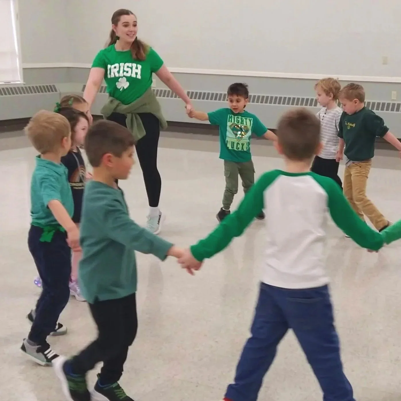 A woman and children holding hands in a circle, participating in a group activity in an indoor room with neutral walls.