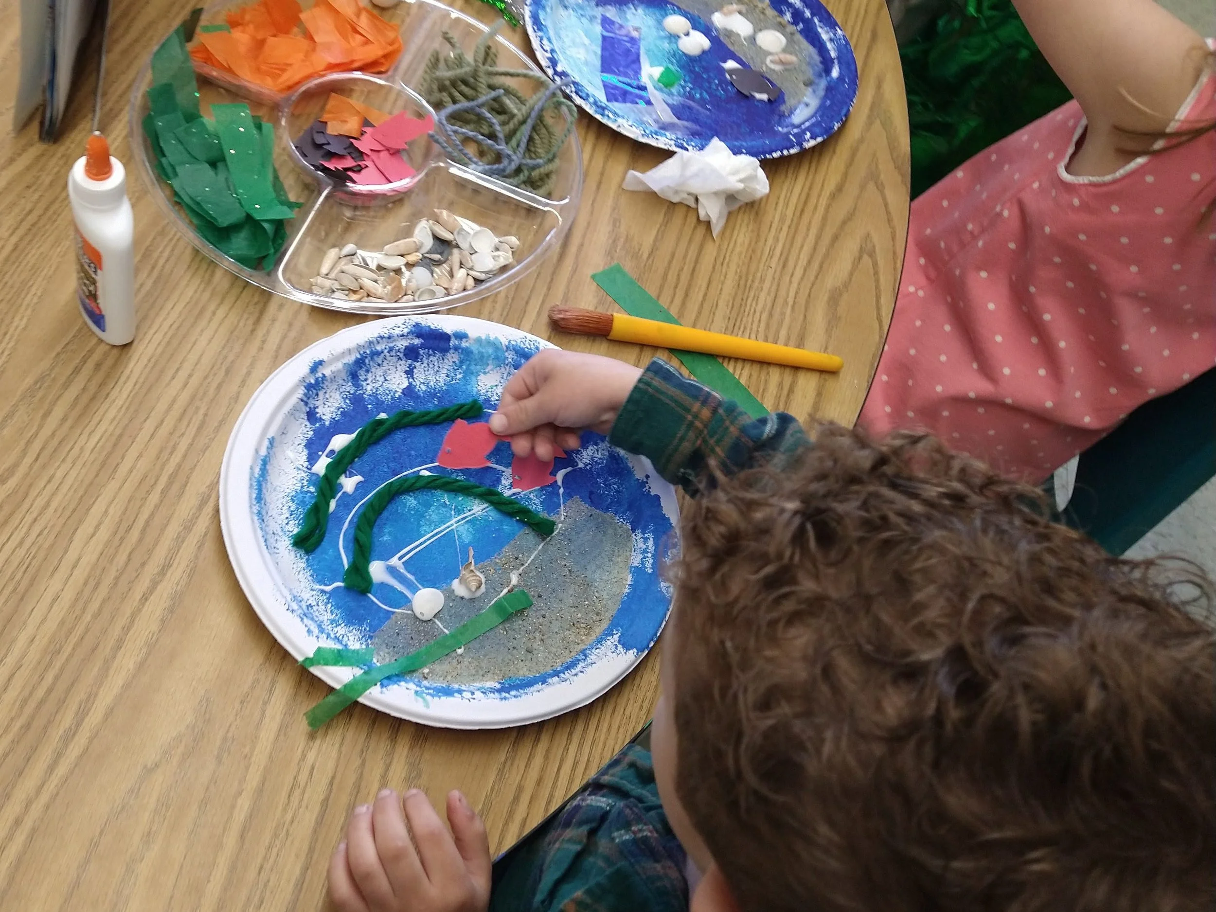 Child engaging in an art activity at a wooden table, with supplies including colored paper, glue, a paintbrush, shells, and painted paper plates with paint and decorations.